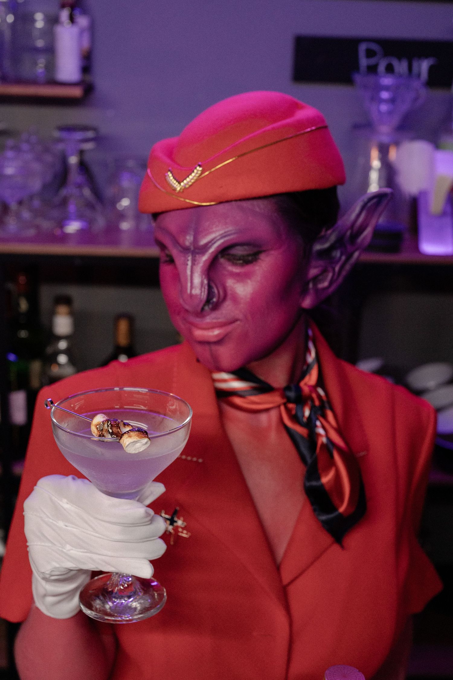 Purple alien makeup artist in red uniform examining cocktail in martini glass at bar.