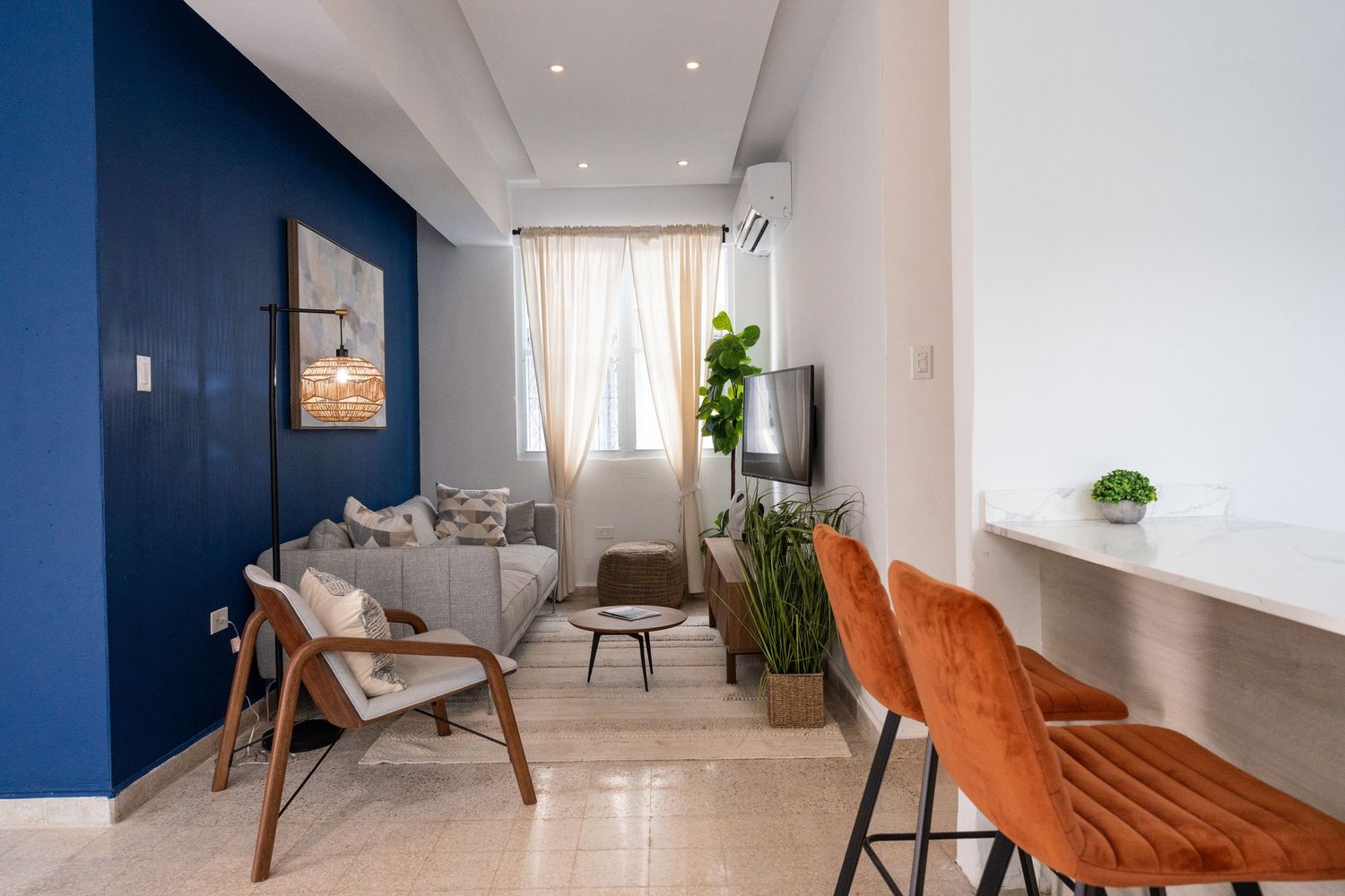 Modern apartment living room with bold blue accent wall, wooden chair, and orange bar stools.
