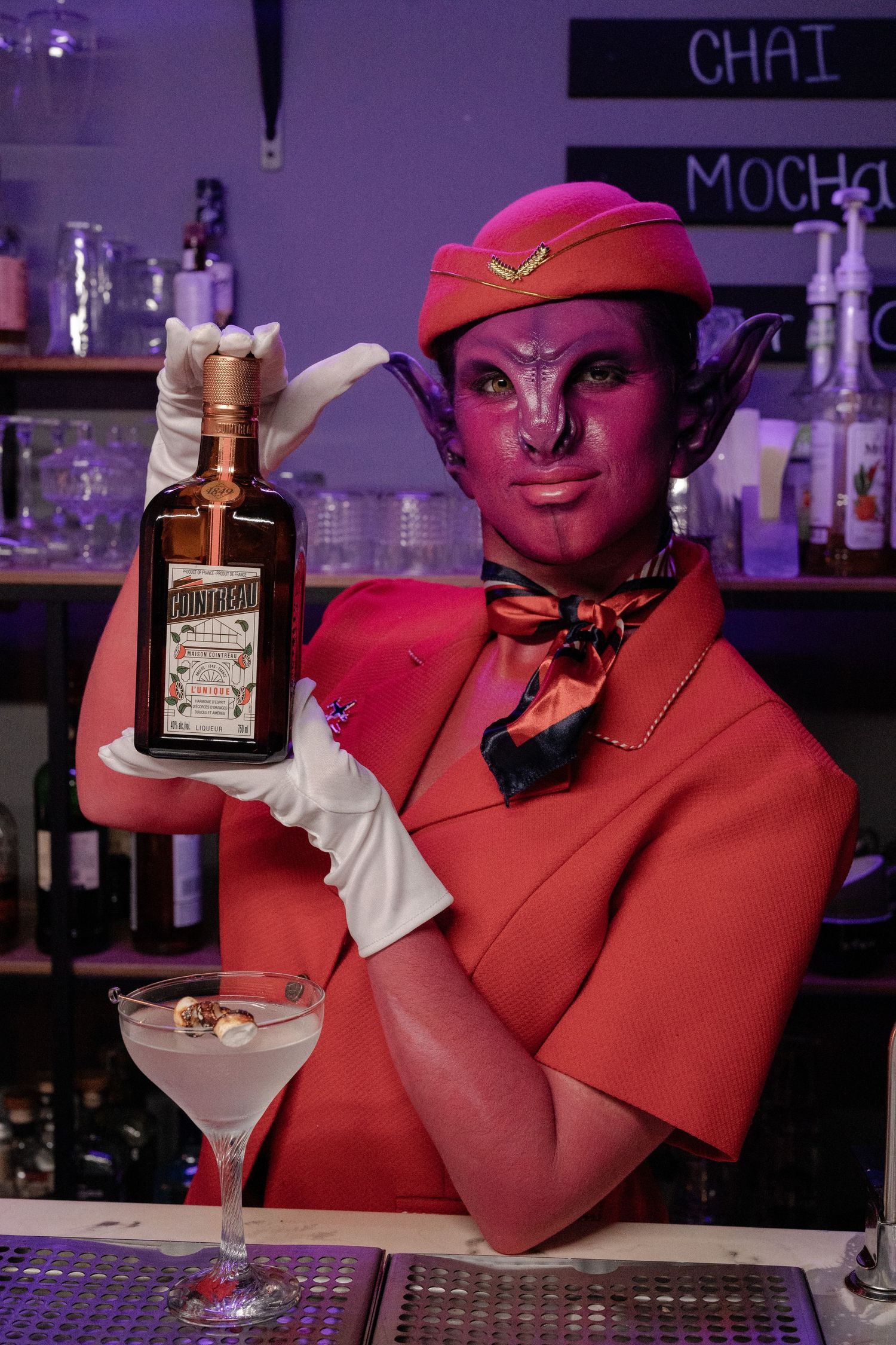 Purple demon character in red flight attendant costume holding Cointreau bottle at a bar.