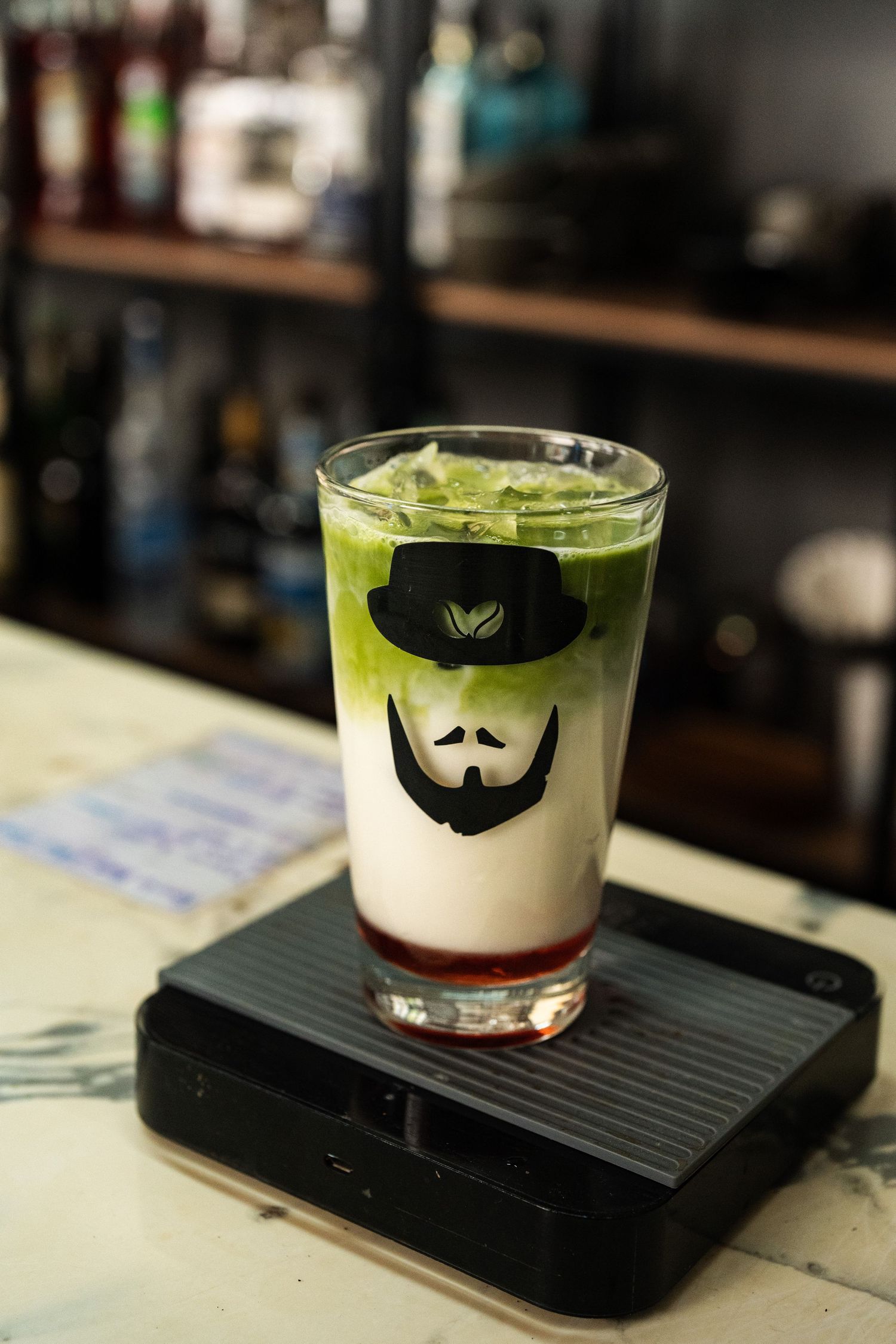 Iced matcha latte in a branded glass with a bearded man logo, layered green and white, on a bar scale.