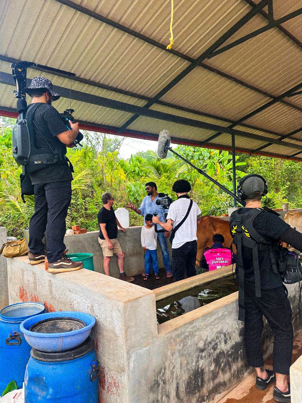 Outdoor film crew shoots an interview scene near a fish pond under a covered structure.