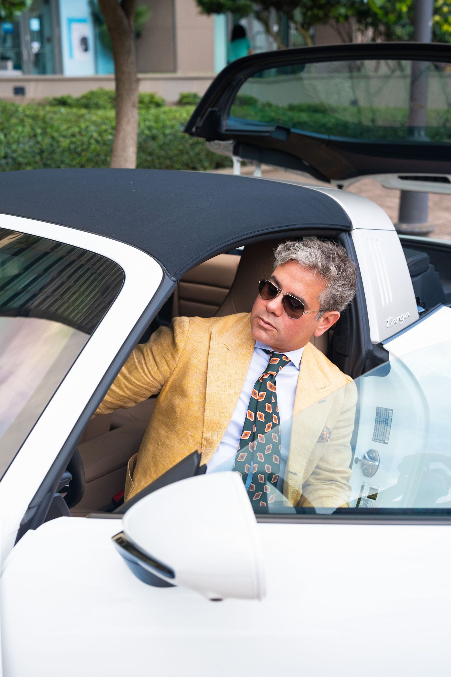 A stylish middle-aged man in a yellow blazer and patterned tie sits in a white convertible Porsche.