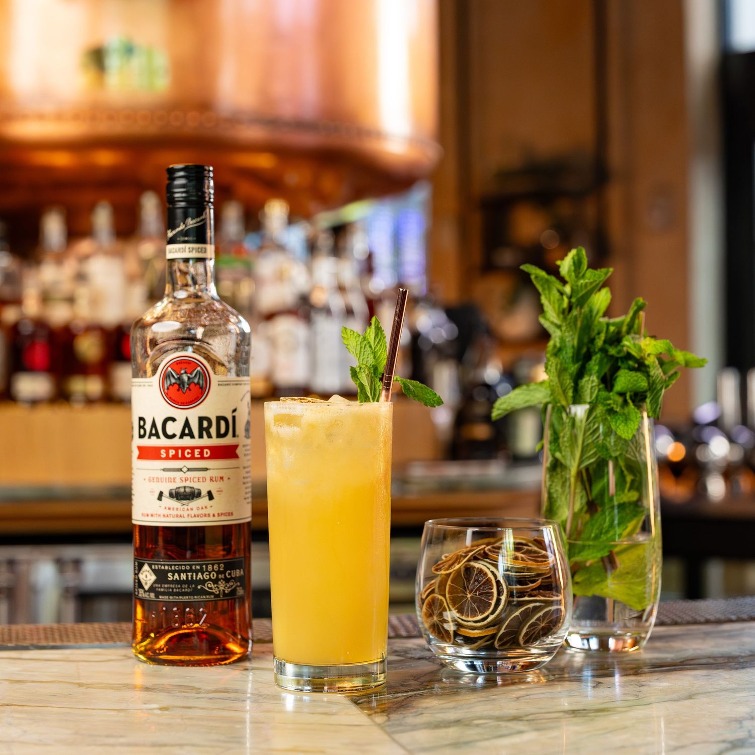Bacardi Spiced rum bottle beside a tropical cocktail, fresh mint garnish, and citrus on a bar counter.