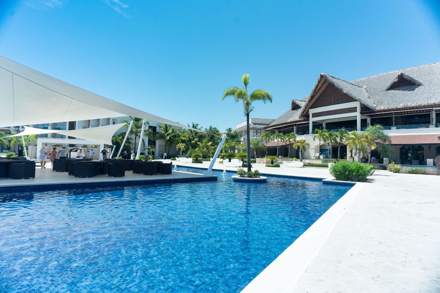 Luxury resort pool with palm trees, white shade sails, and tropical architecture on sunny day.