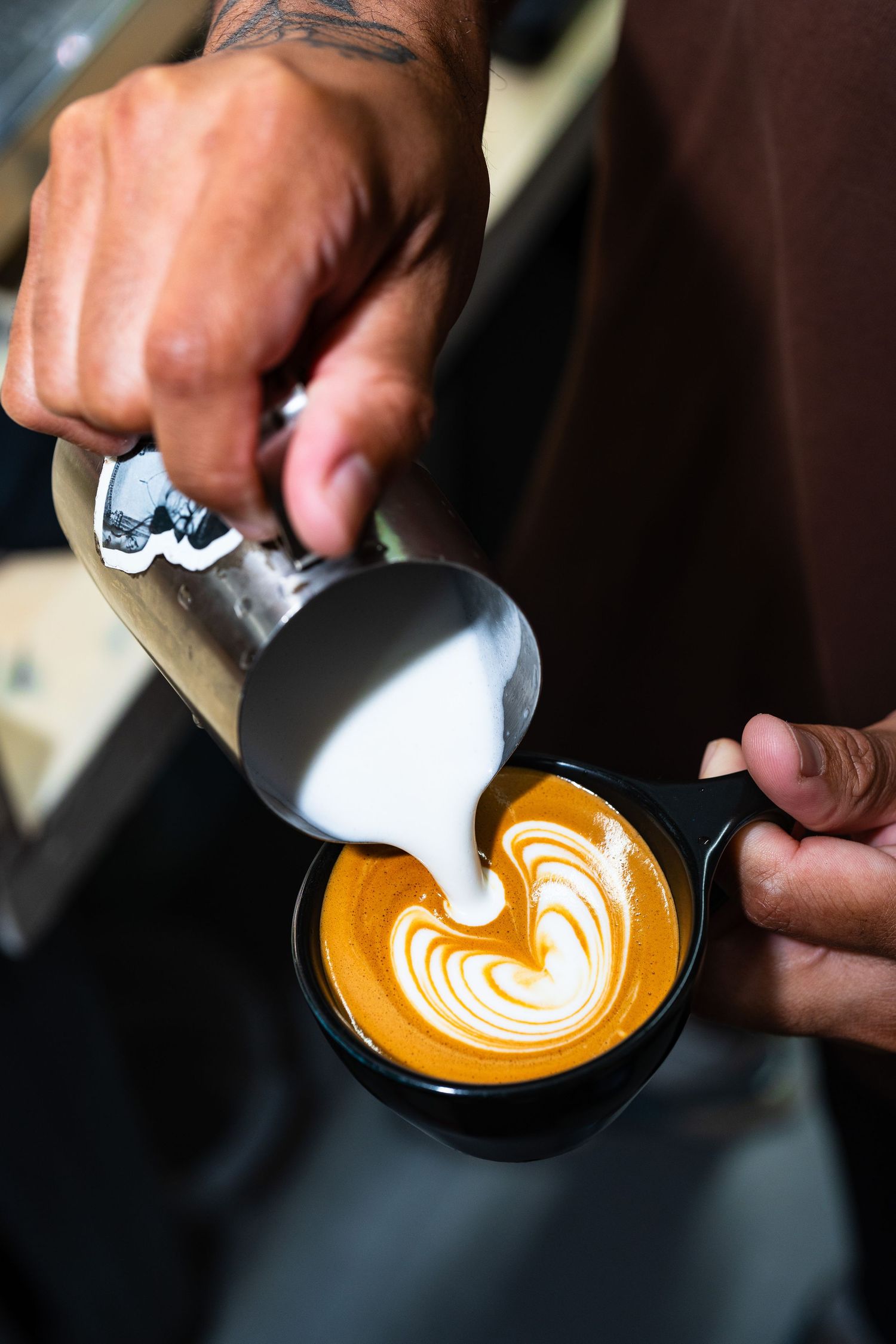 Skilled barista pours steamed milk creating heart latte art in a black coffee cup.