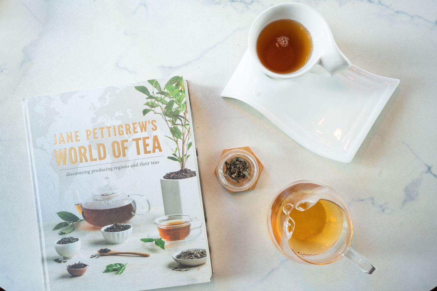 Overhead view of Jane Pettigrew's World of Tea book beside a cup of tea and loose leaf tea on marble.
