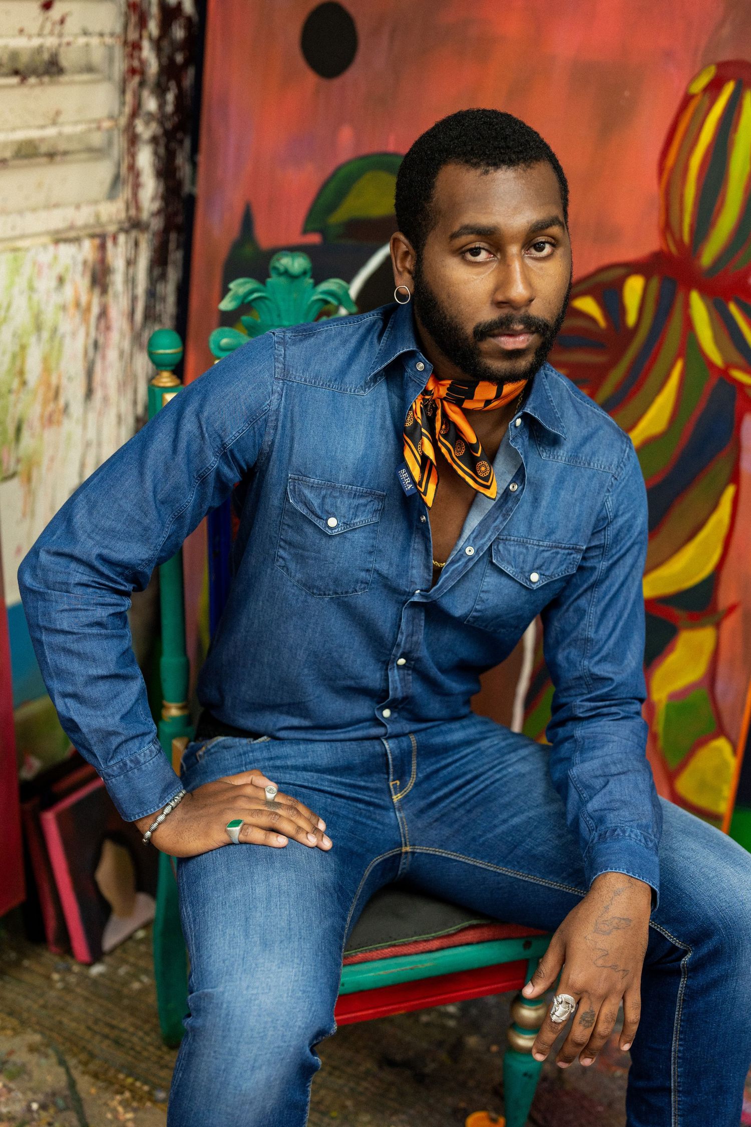 Stylish Black man in denim outfit with orange scarf sitting before colorful graffiti wall backdrop.