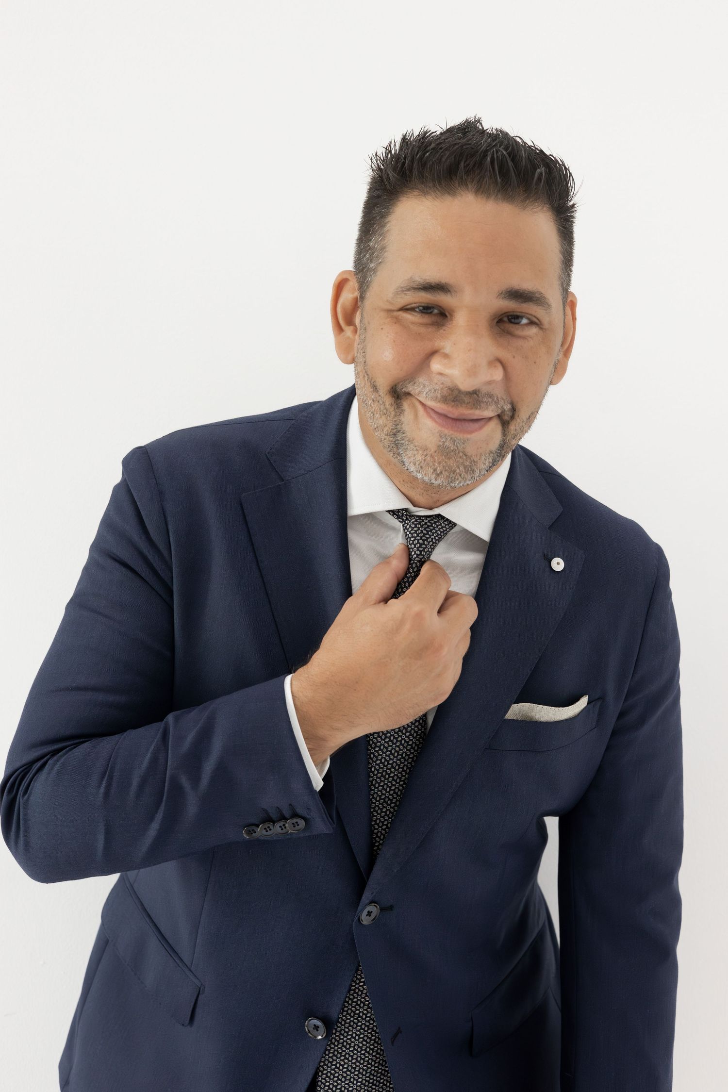 Cheerful man in navy suit adjusting his patterned tie against white background.