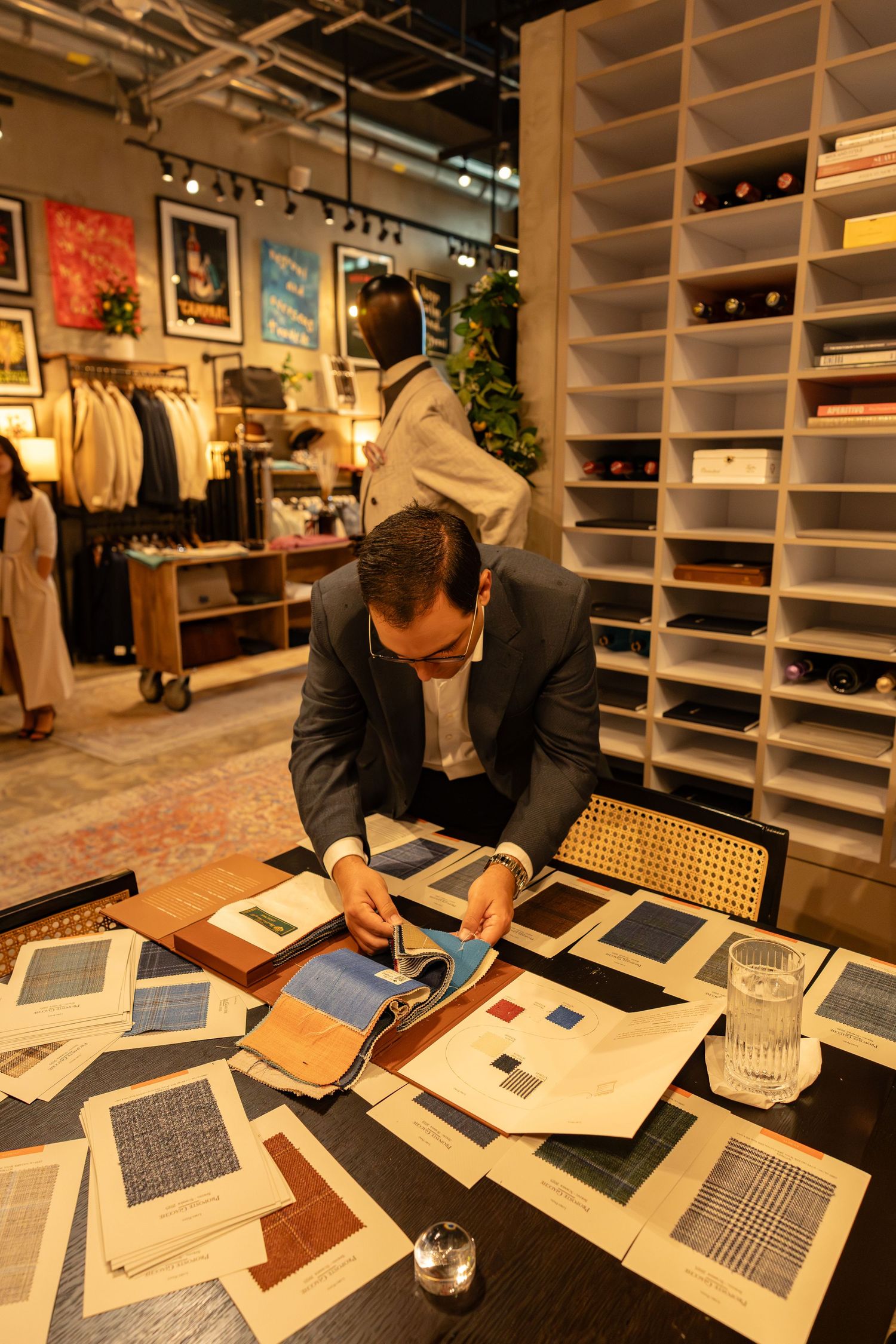 Man in suit examines fabric swatches spread across a table in a modern menswear boutique.