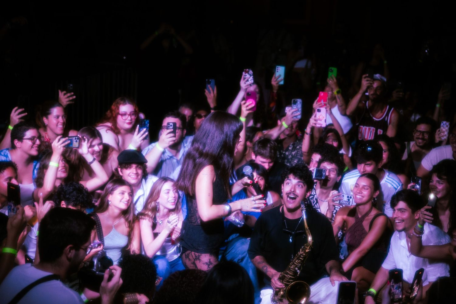 Energetic crowd at a live concert with a saxophonist performing among fans under colorful stage lighting.