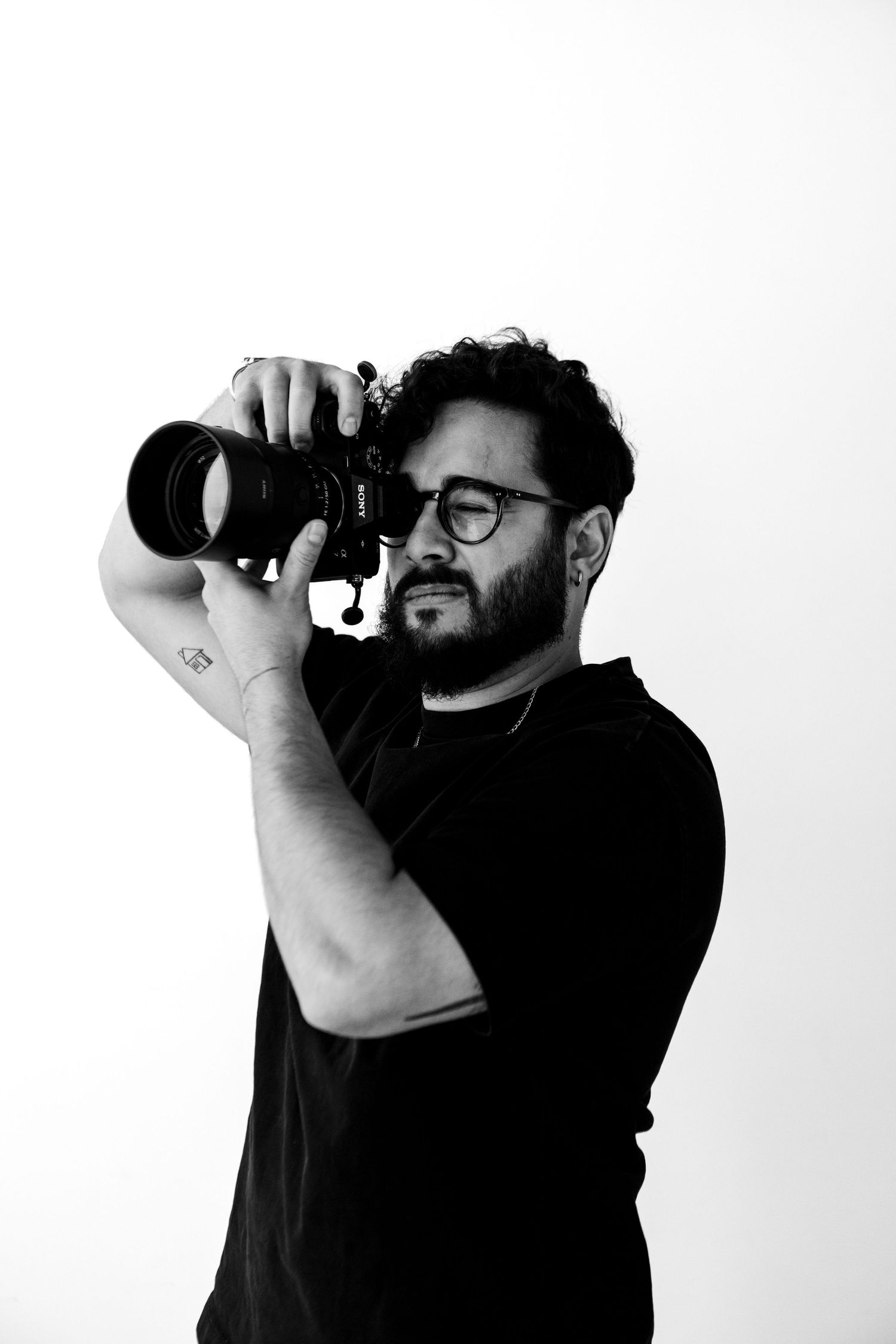 Black and white portrait of a bearded man with glasses holding a camera up to his eye against white background.