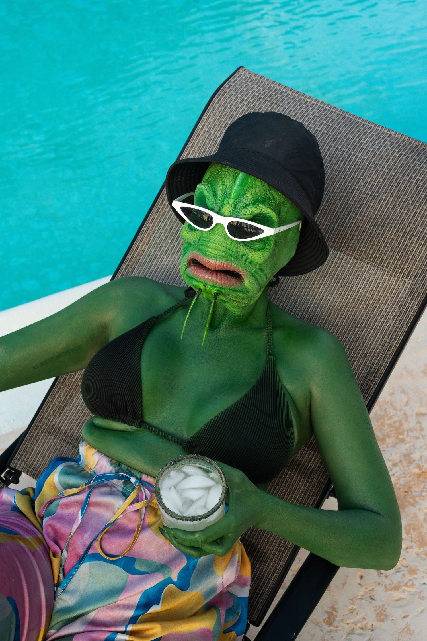 Person in green fish monster makeup relaxing on pool lounger with sunglasses and cocktail.