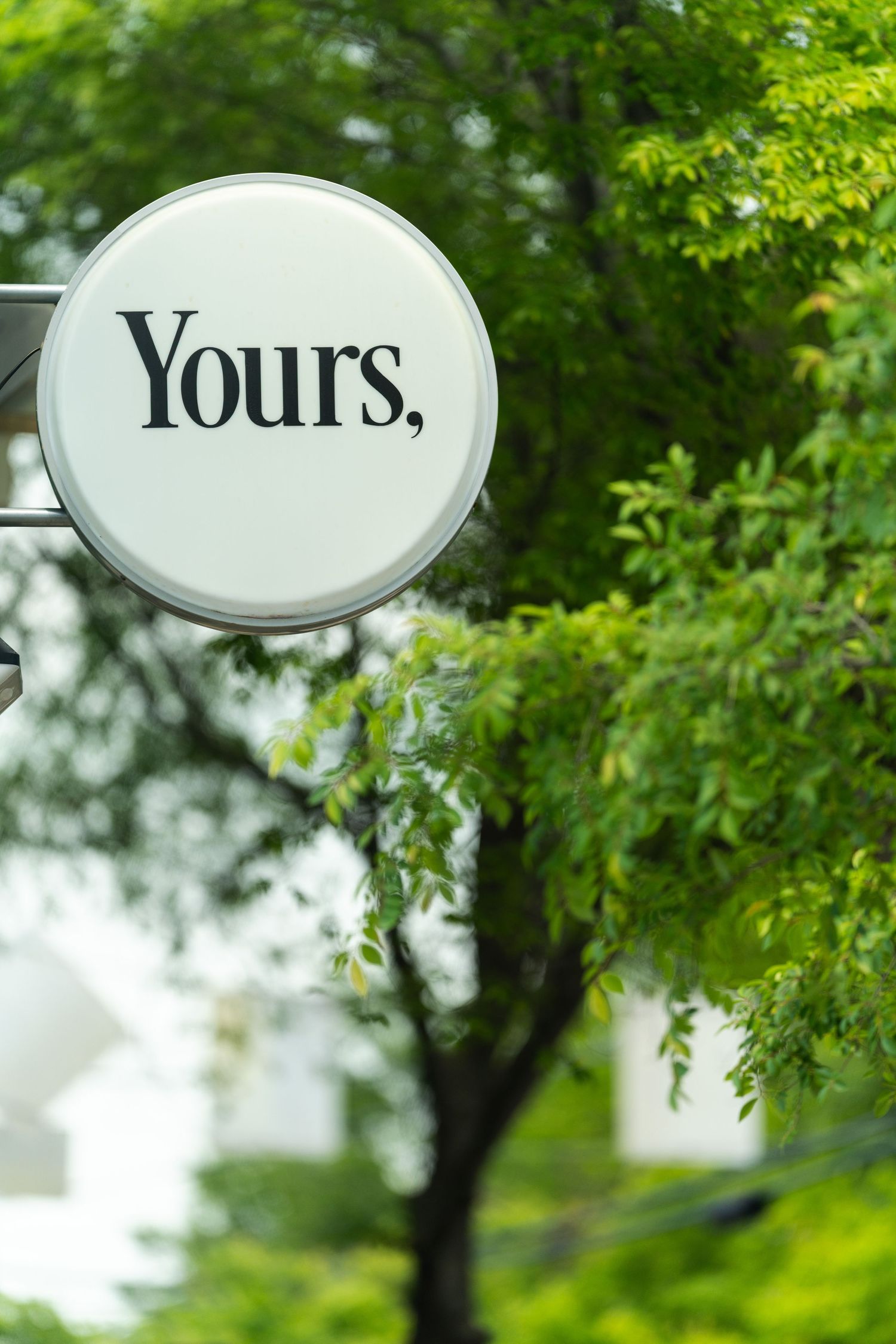 Round white outdoor sign reading Yours, mounted on a post surrounded by green trees.