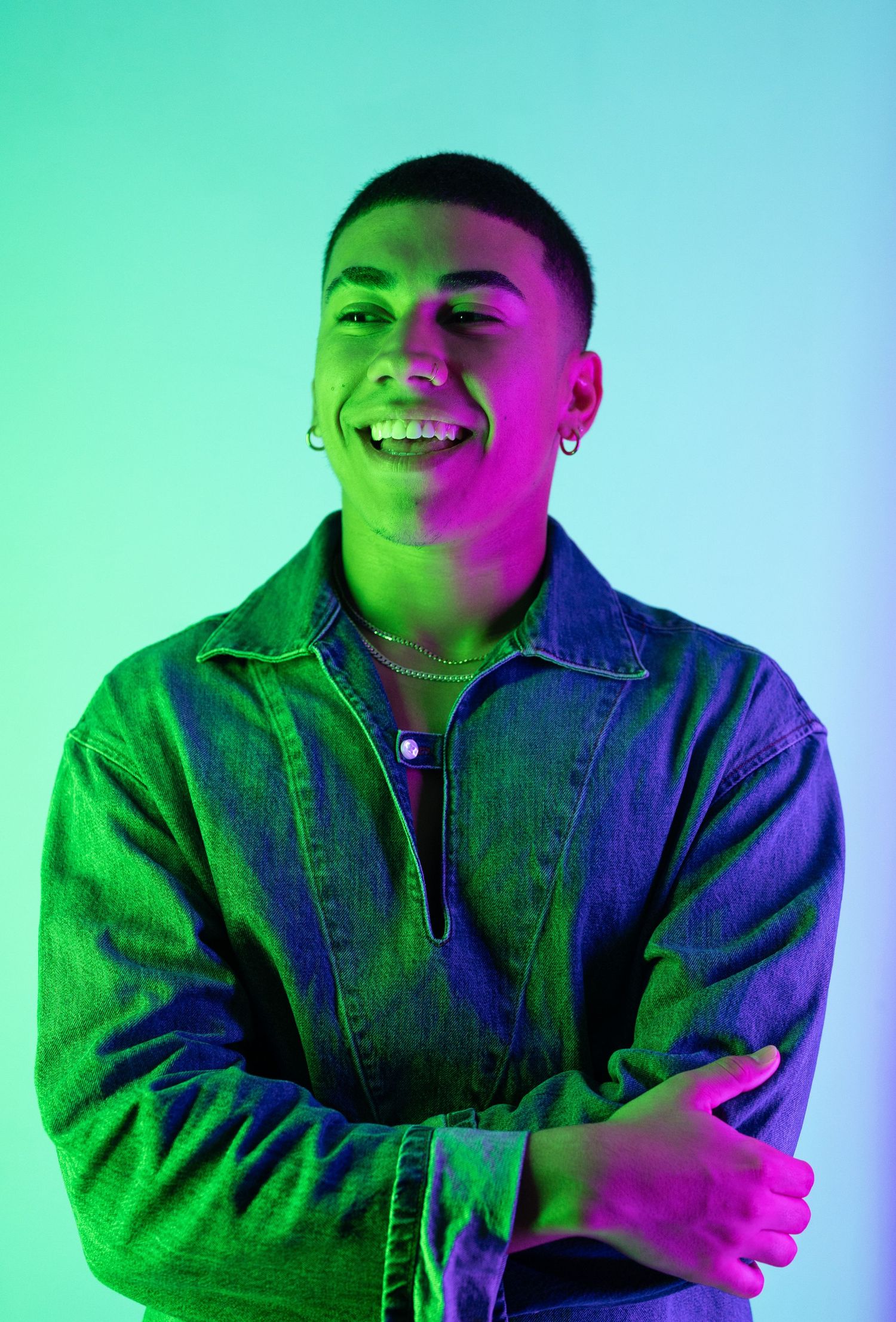 Smiling young man in denim jacket with crossed arms under vibrant green and purple neon lights.