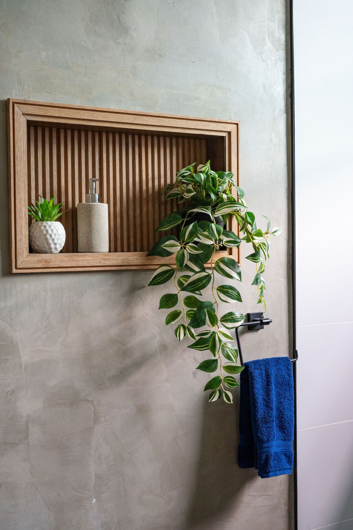 Wooden niche shelf with plant, soap dispenser, and blue towel in modern bathroom.