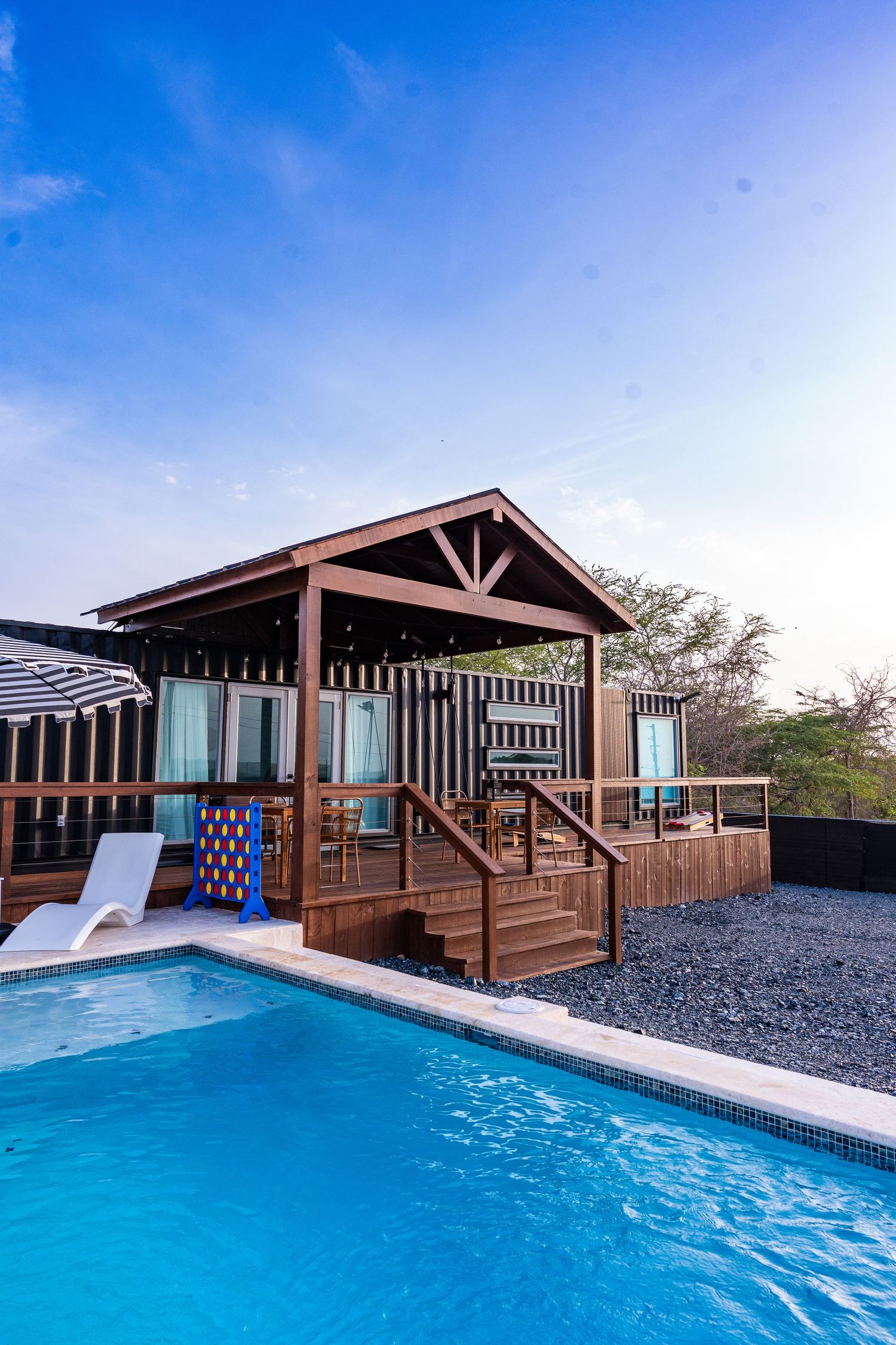 Wooden pool cabana with blue swimming pool under a bright blue sky at a resort.