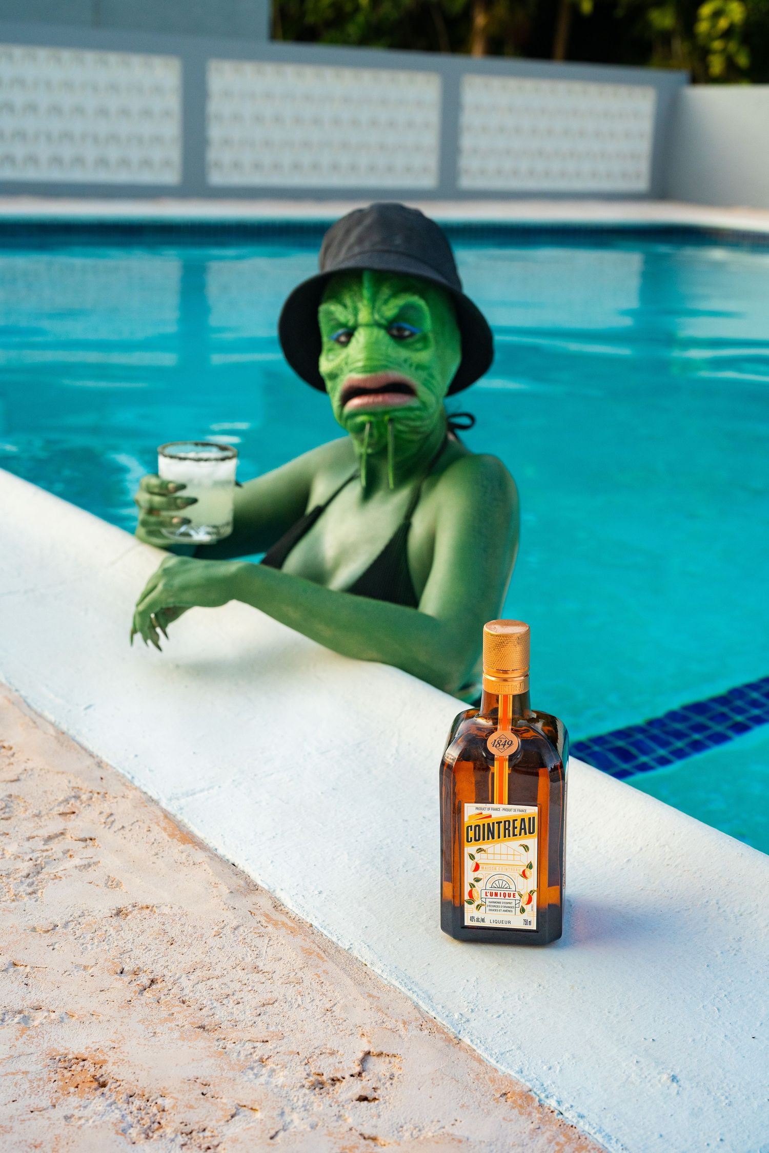 Green sea creature costumed person in pool holding cocktail with Cointreau bottle on ledge.