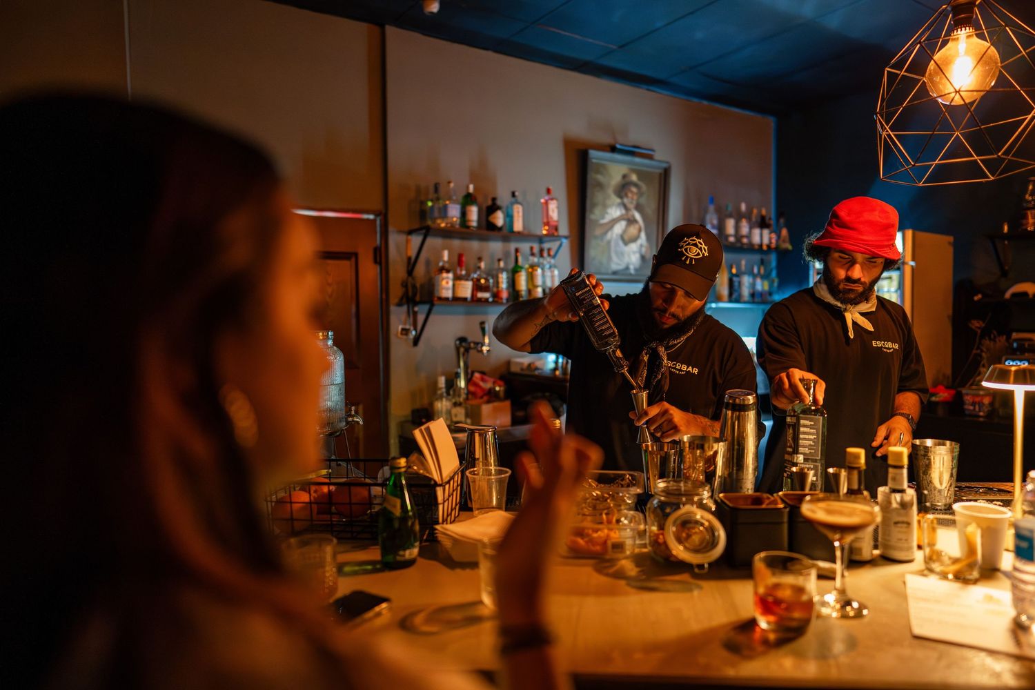 Two bartenders preparing cocktails behind a dimly lit bar while a customer watches from the foreground.