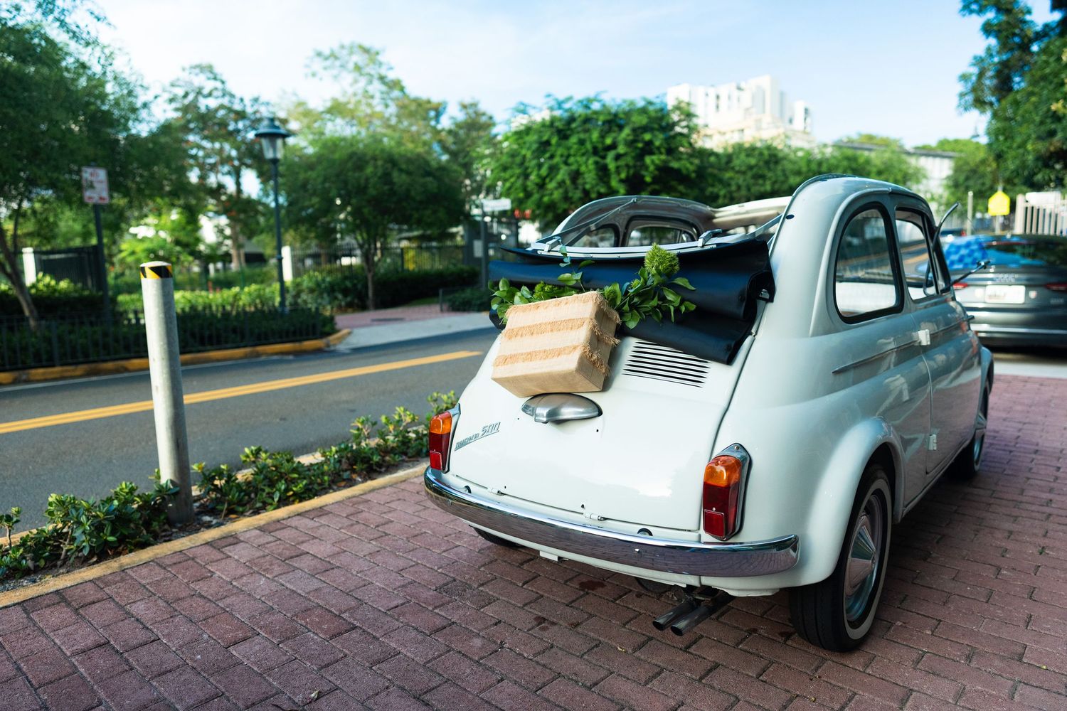 Vintage light blue Fiat 500 with open trunk containing brown box, parked on brick street.