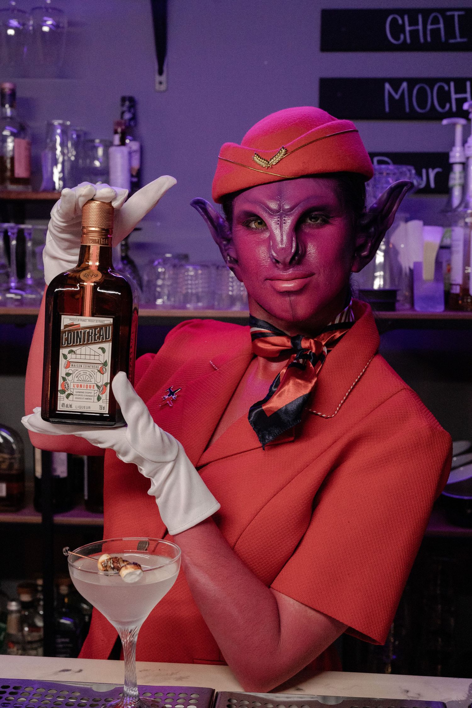 Pink alien cosplayer in red uniform holds Cointreau bottle beside a martini glass at a bar.