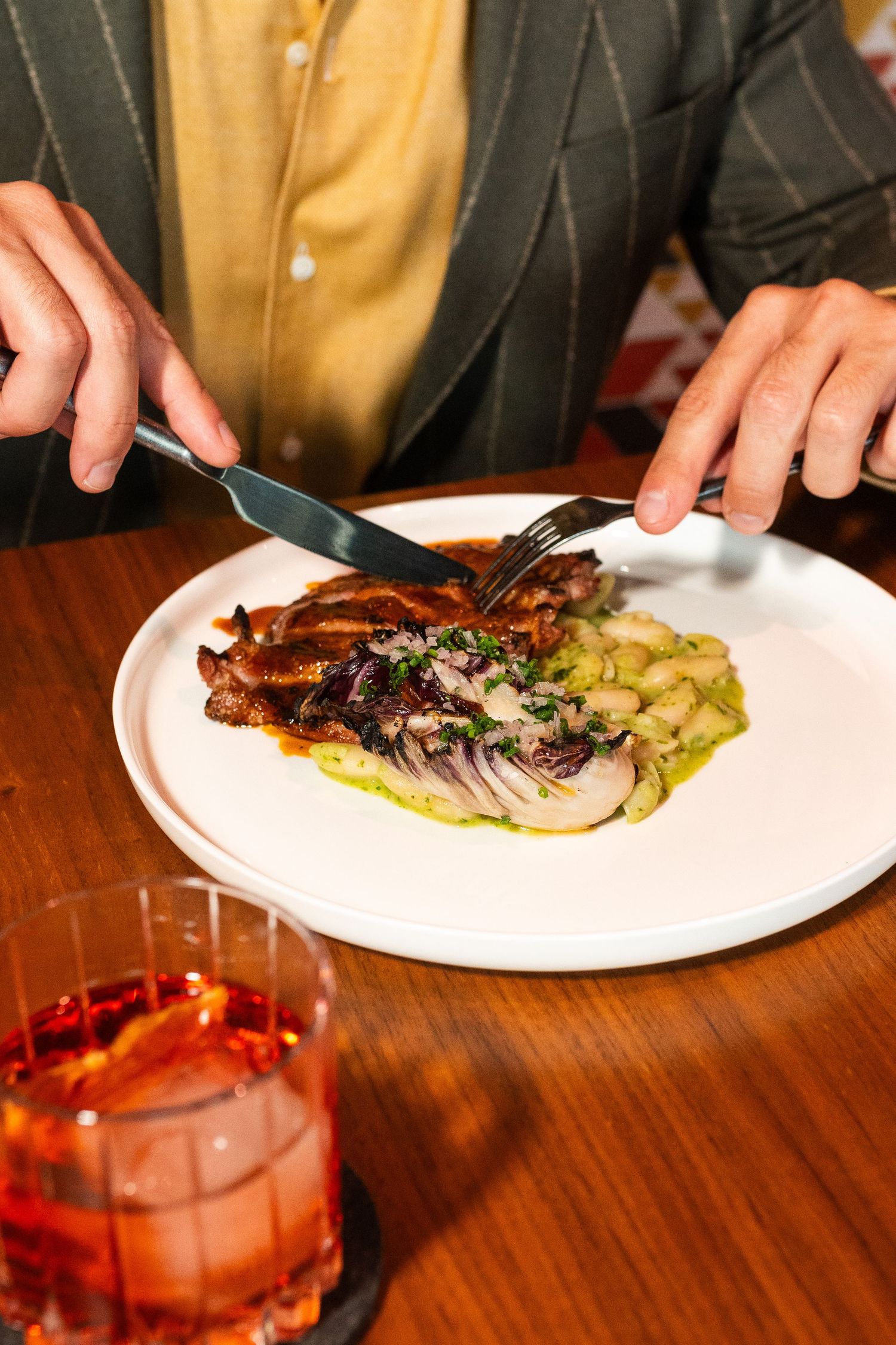 Person cutting grilled meat with white beans and chimichurri on white plate beside red cocktail.