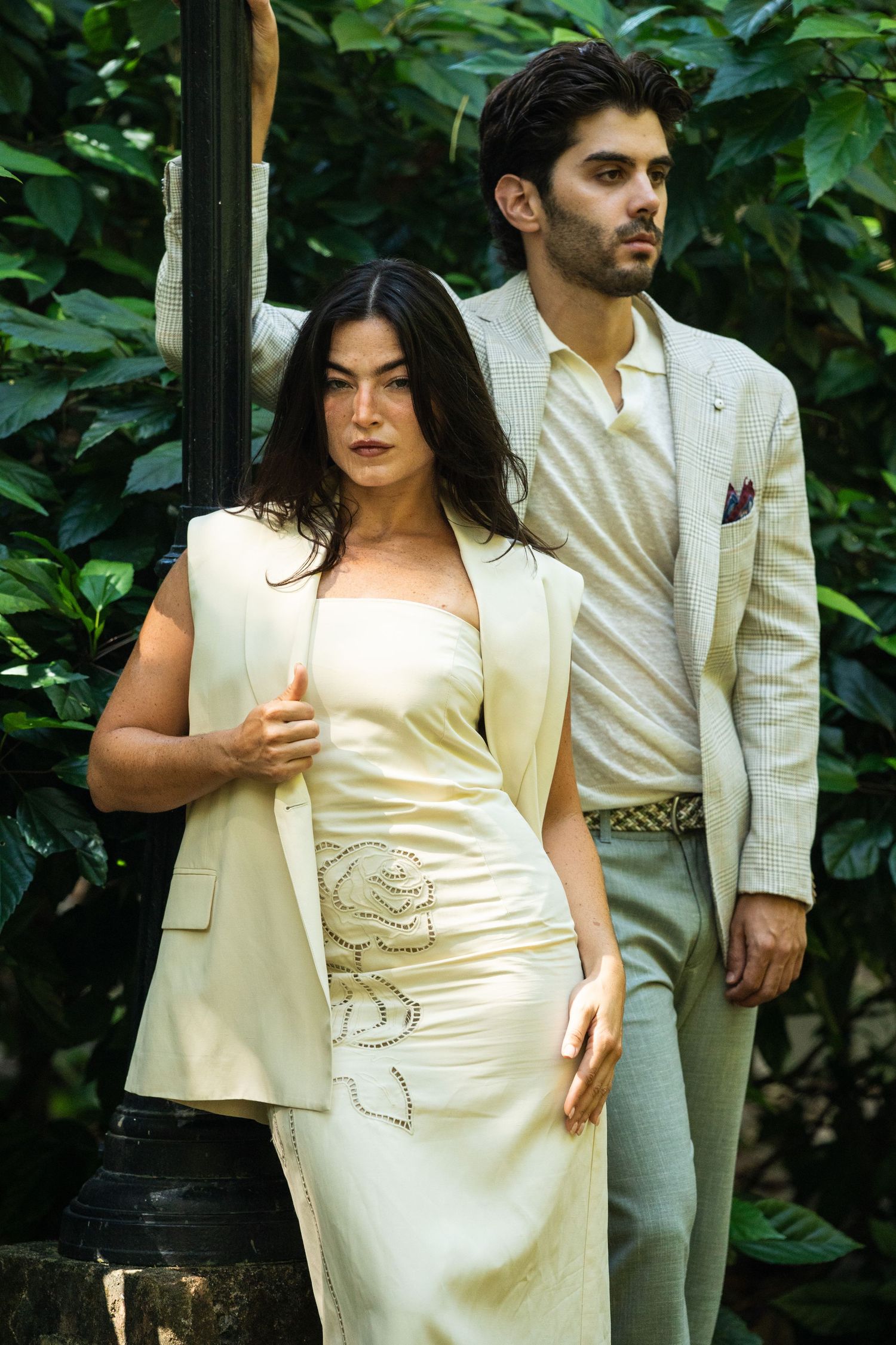 Elegant couple in cream formal attire posing among lush green foliage outdoors.