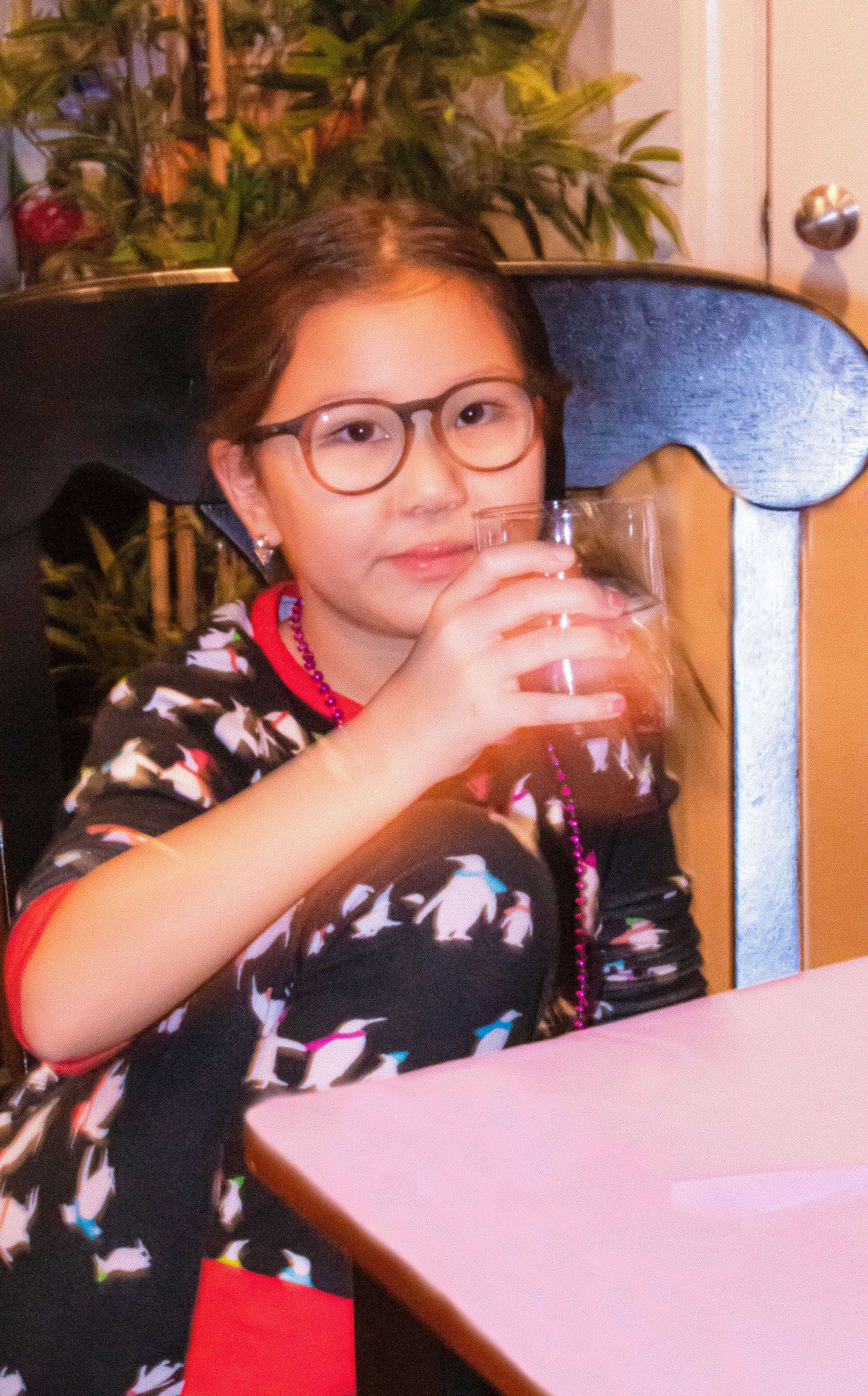 Young person wearing glasses sips from a glass with a festive setting in the background.