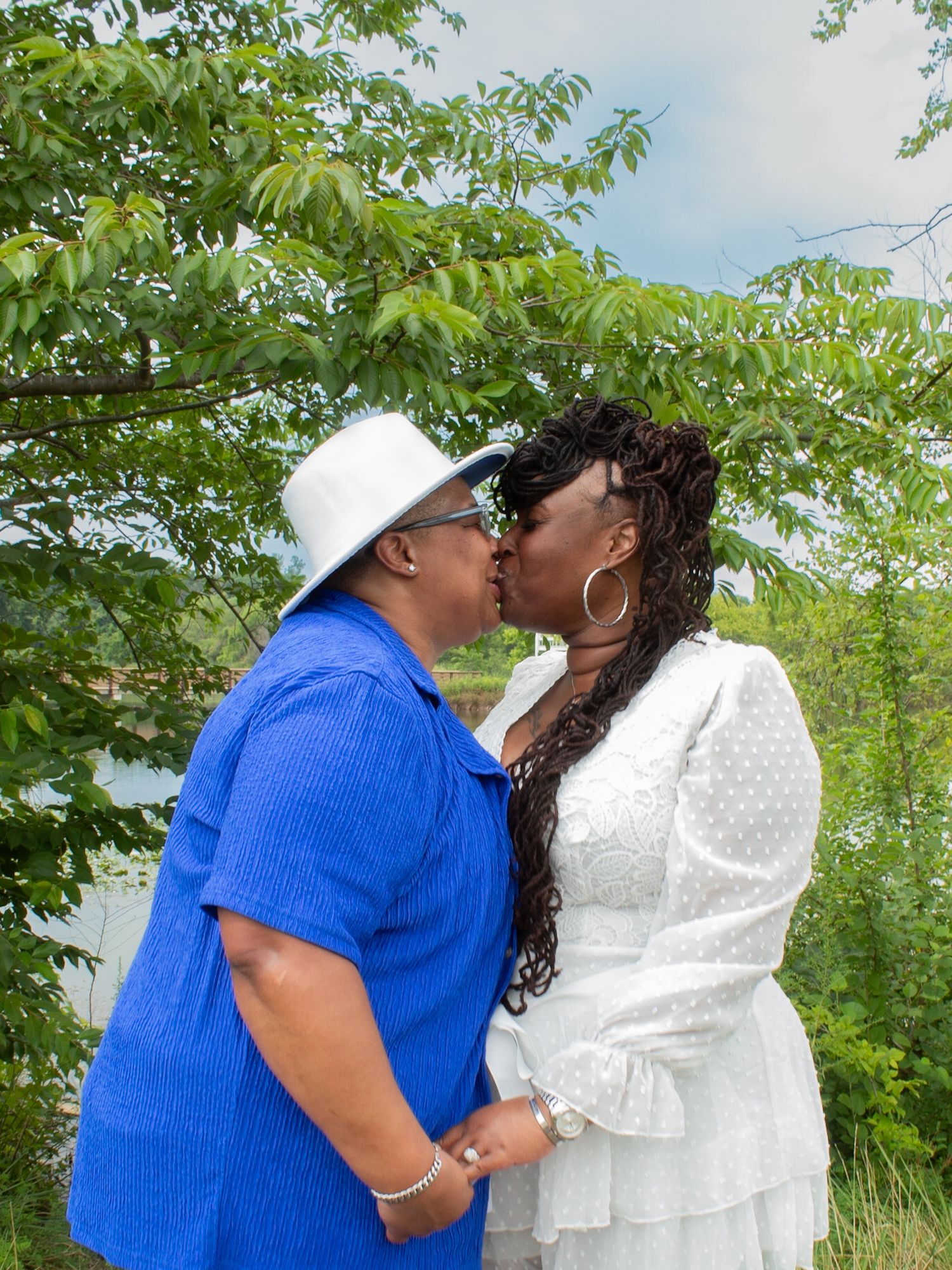 A couple in blue and white outfits sharing an intimate moment outdoors surrounded by green foliage.