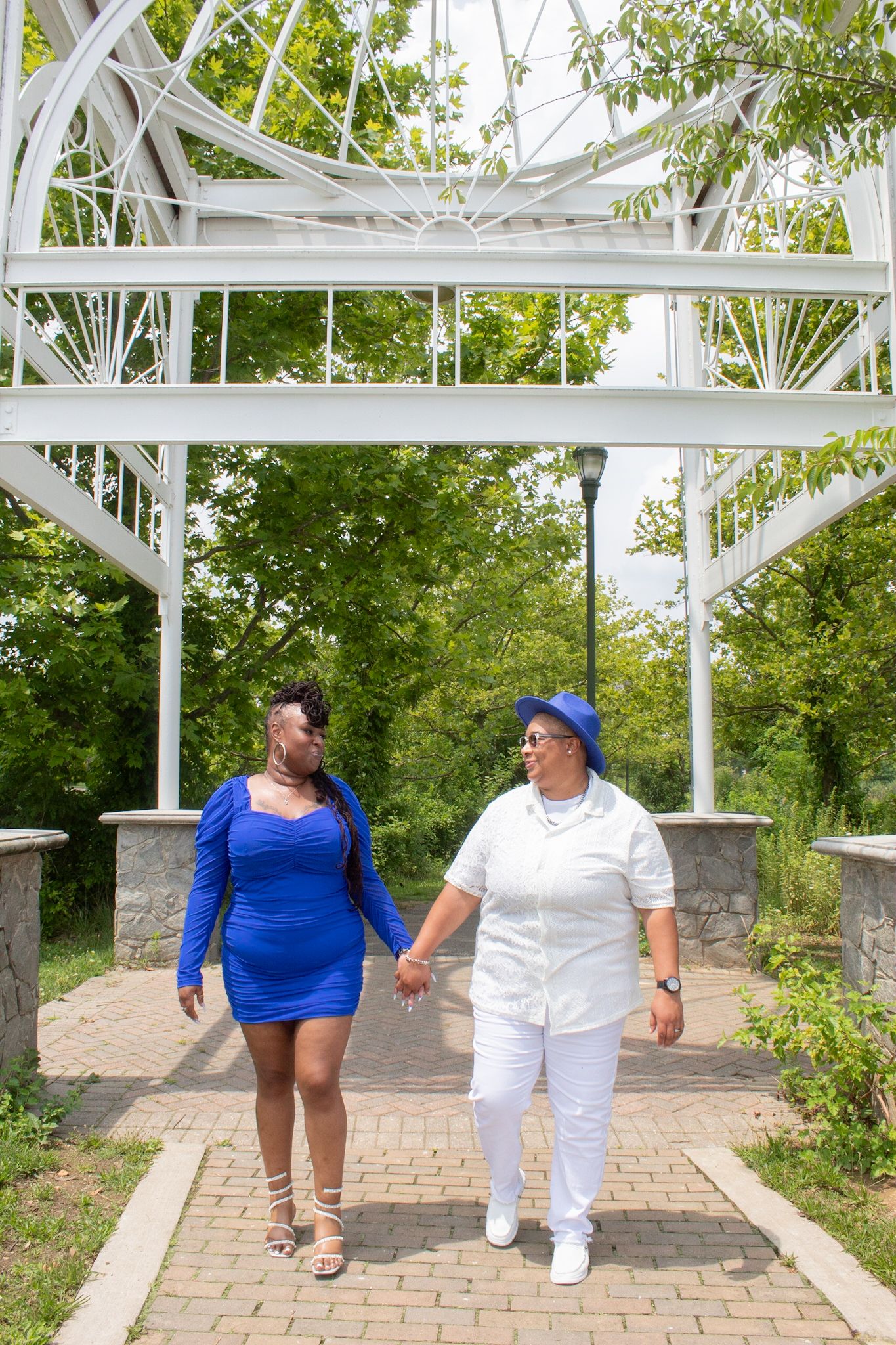 Two people walk along a brick path under a white pergola in a garden setting wearing coordinated blue and white outfits.