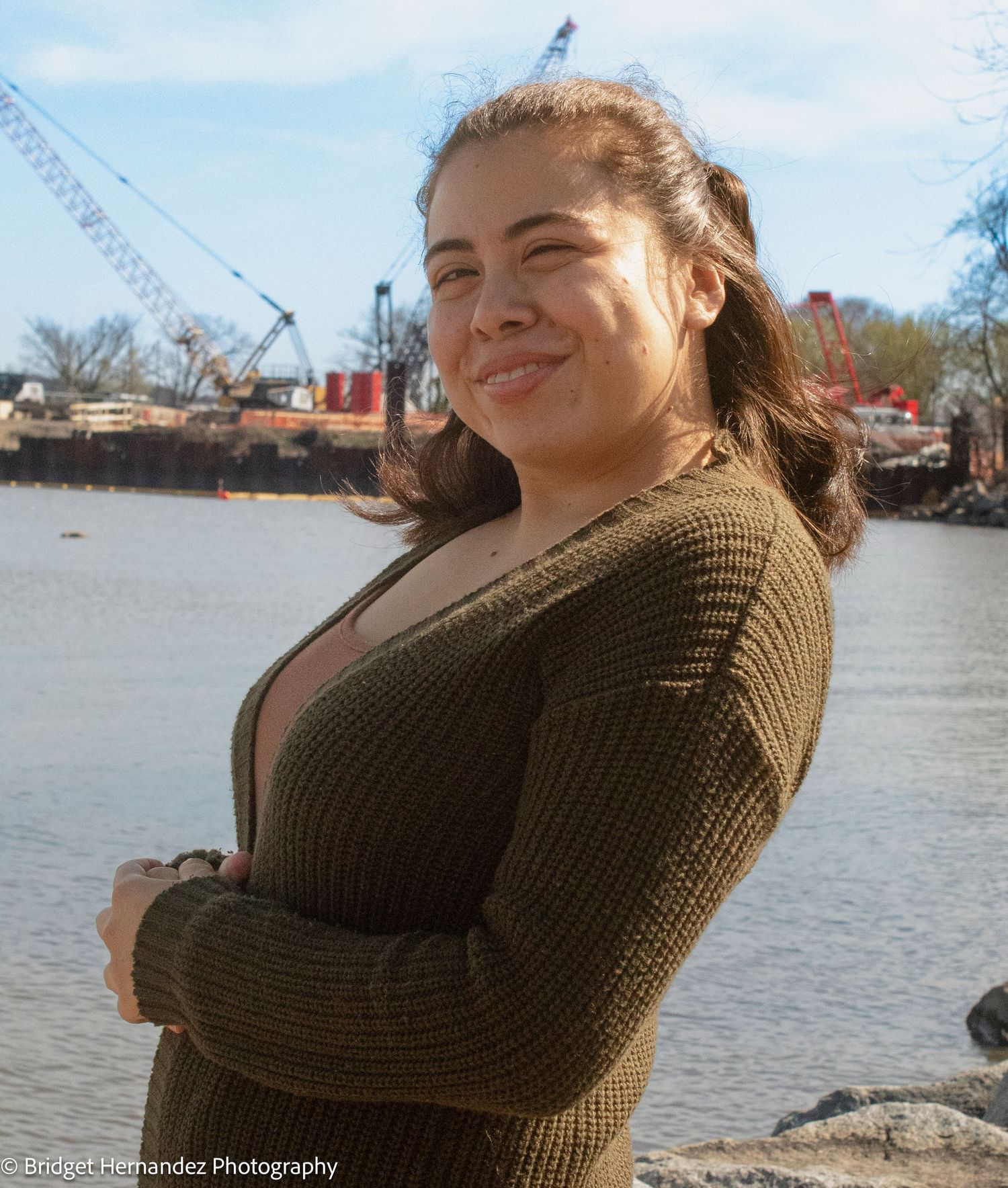 Casual portraits by waterfront wearing brown knit sweater with industrial cranes in background.