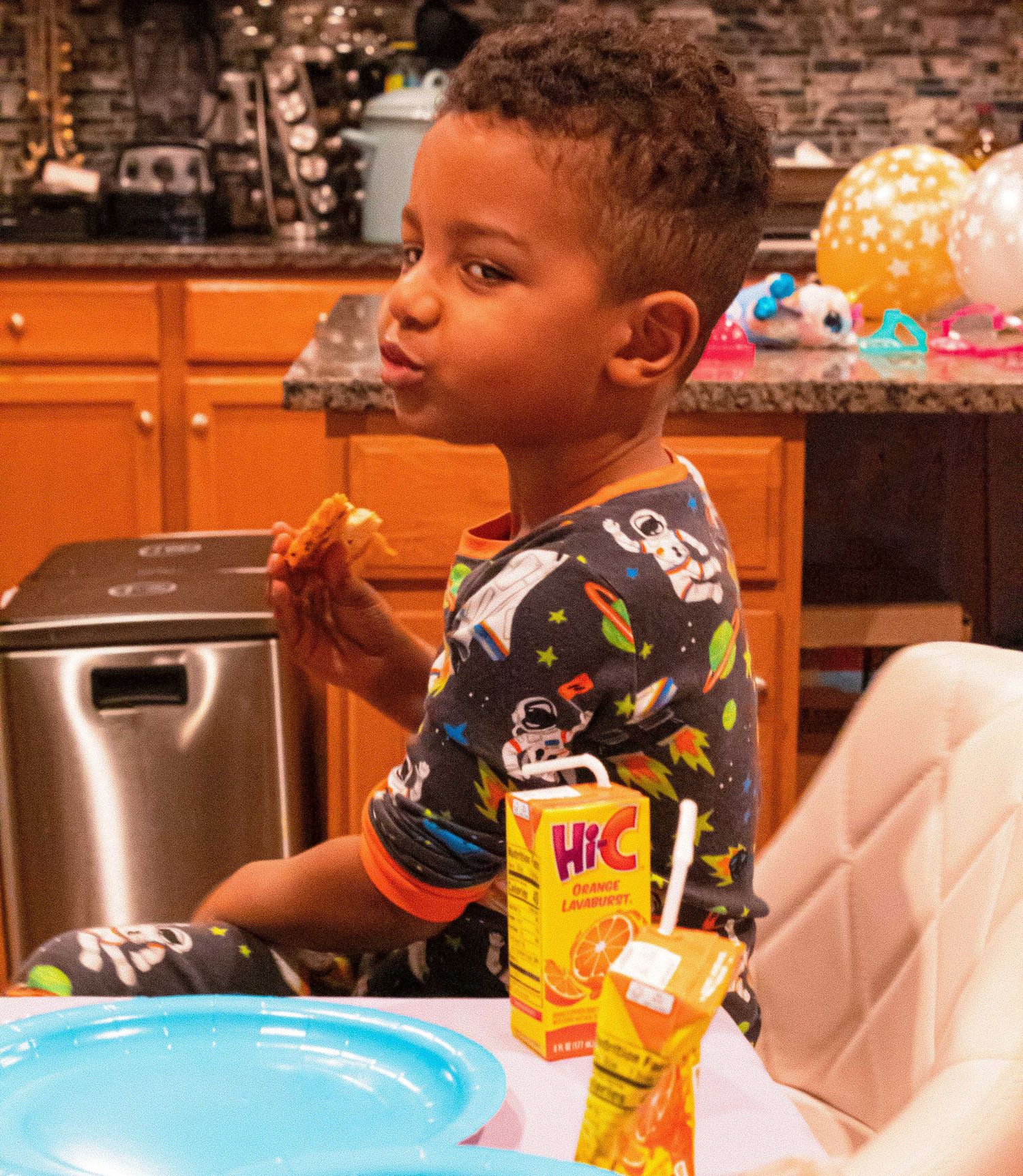 Child drinking from a juice box at a table with party snacks and decorations visible.