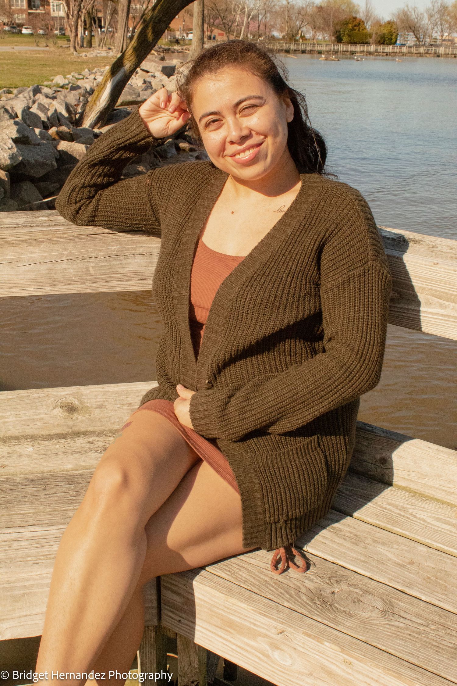 Person in brown cardigan relaxing on wooden dock by waterfront on sunny day.