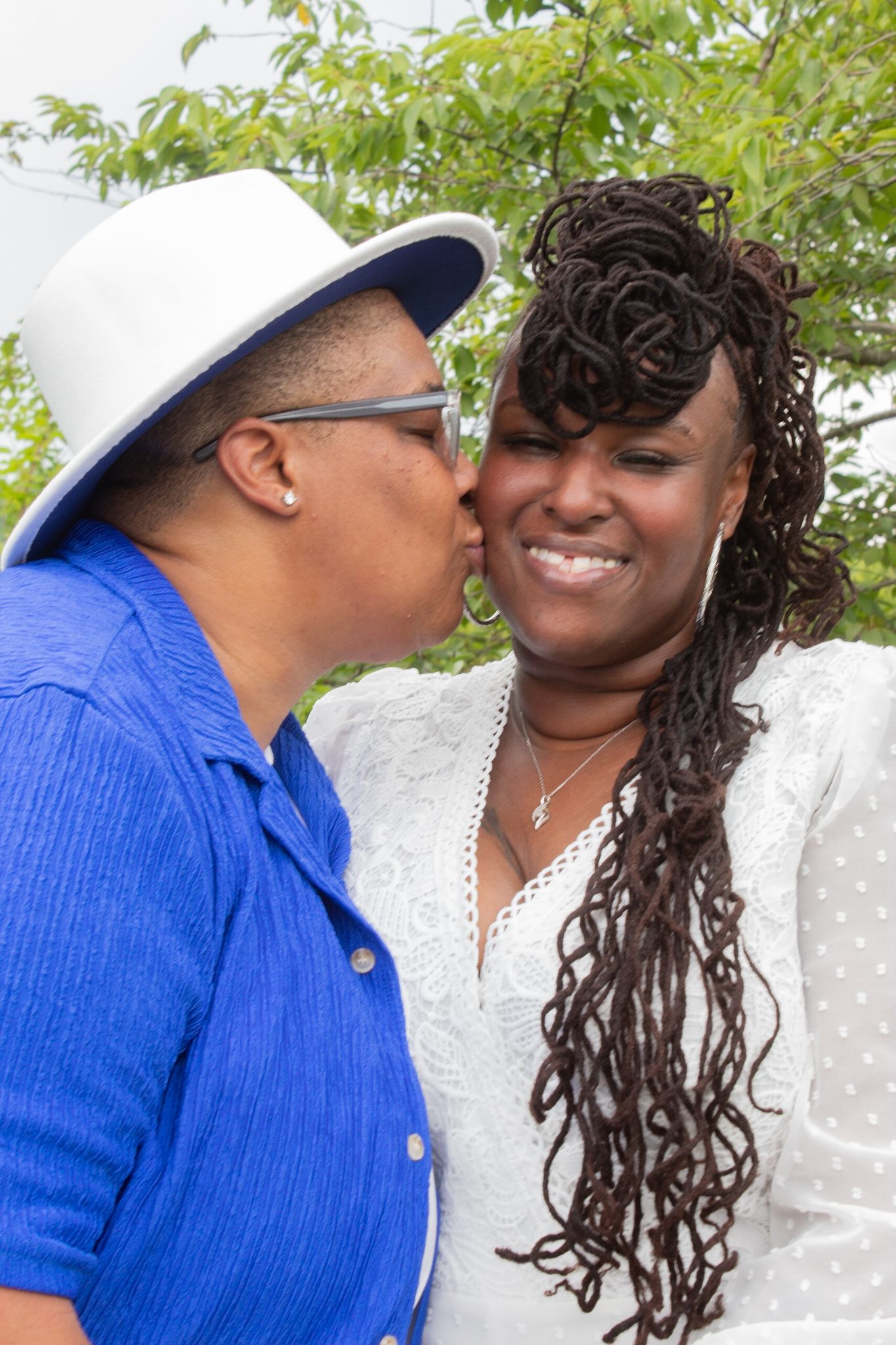 Two people in contrasting blue and white attire sharing a tender moment against a leafy backdrop.
