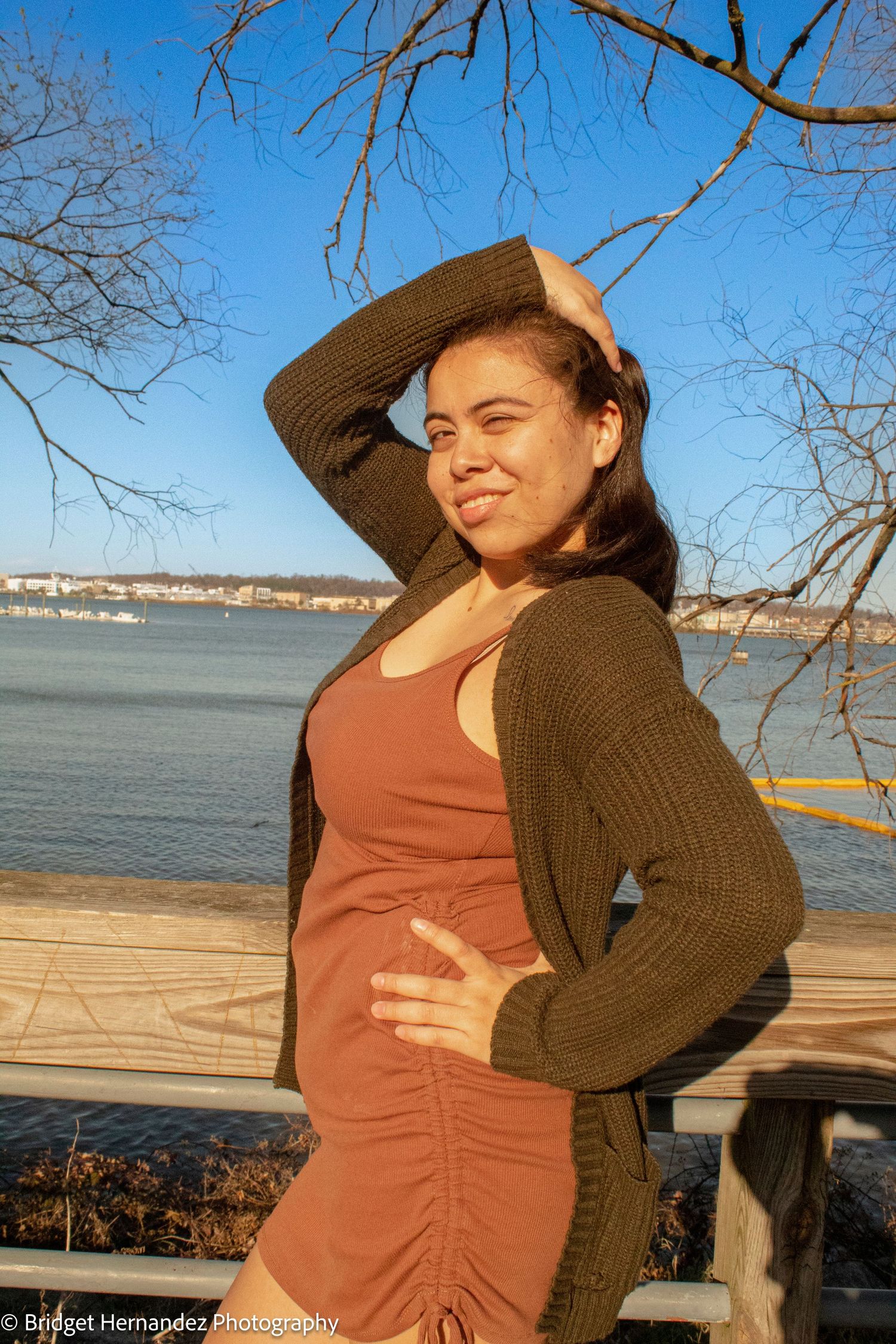 Woman in brown sweater and orange dress poses playfully by waterfront on sunny day.