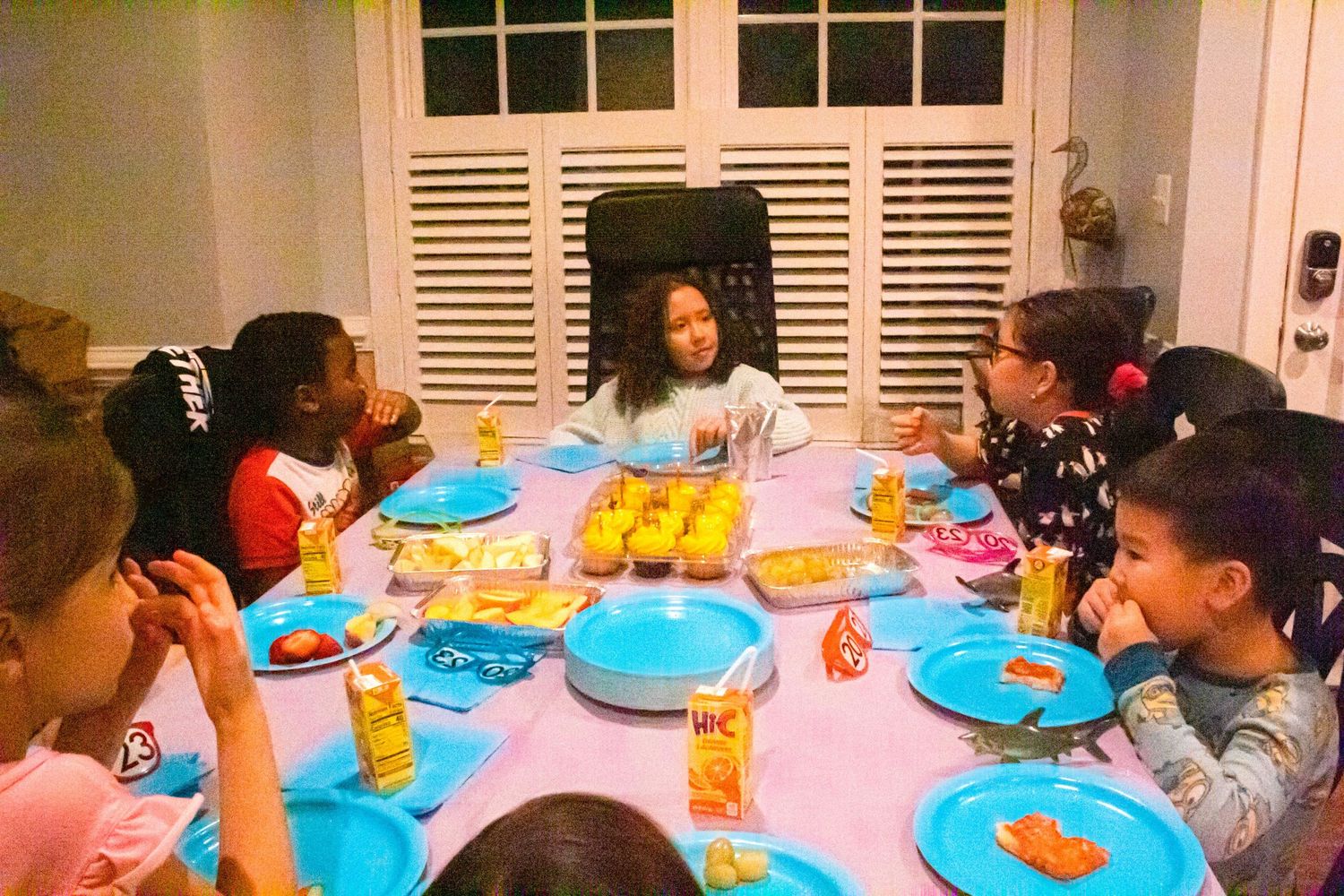 Children seated at a party table with blue plates and pink tablecloth enjoying snacks.