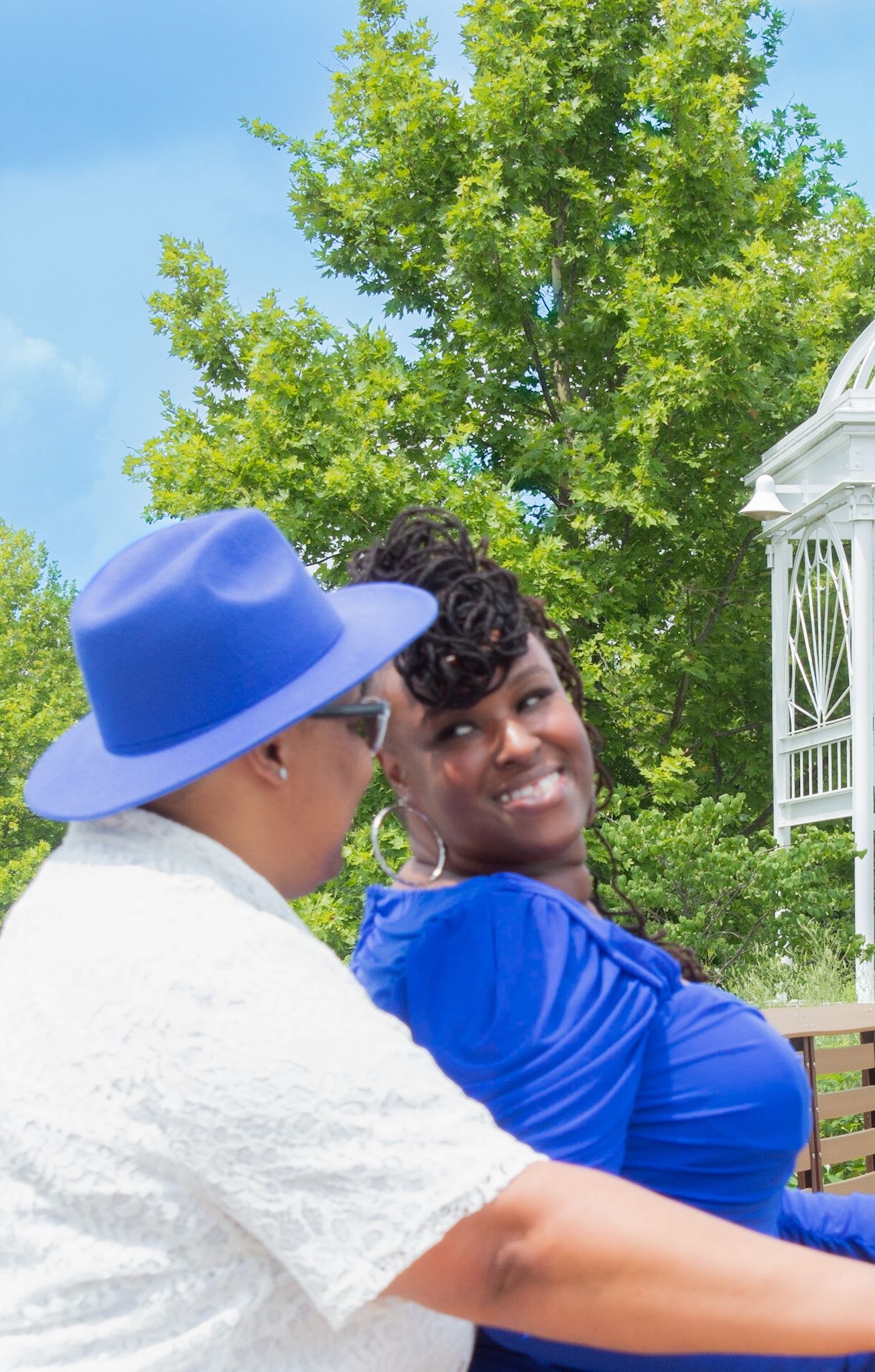 Couple poses together on wooden boardwalk near decorative white gazebo on sunny afternoon.
