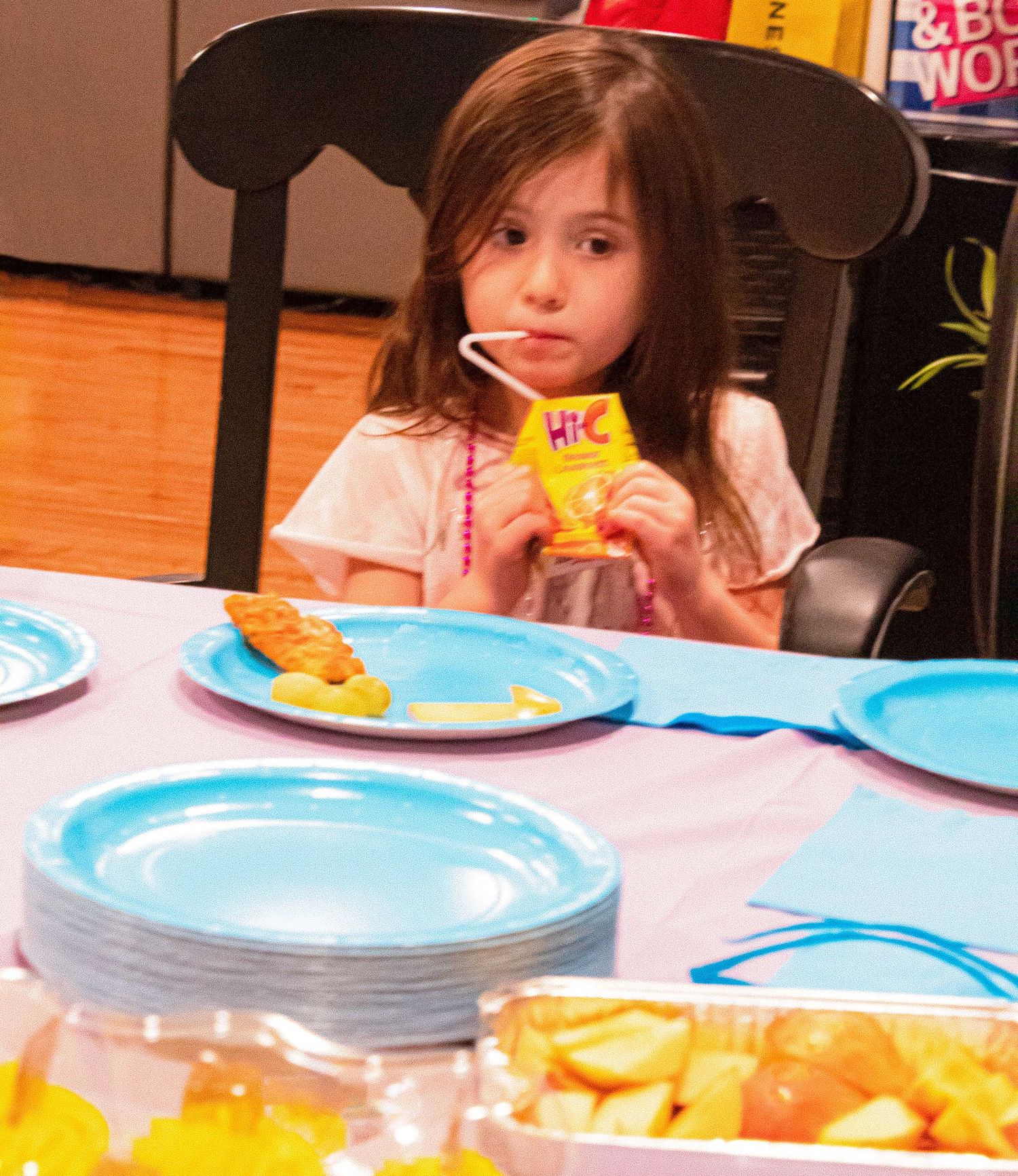 A young party guest sips a yellow drink while sitting at a table with blue plates.