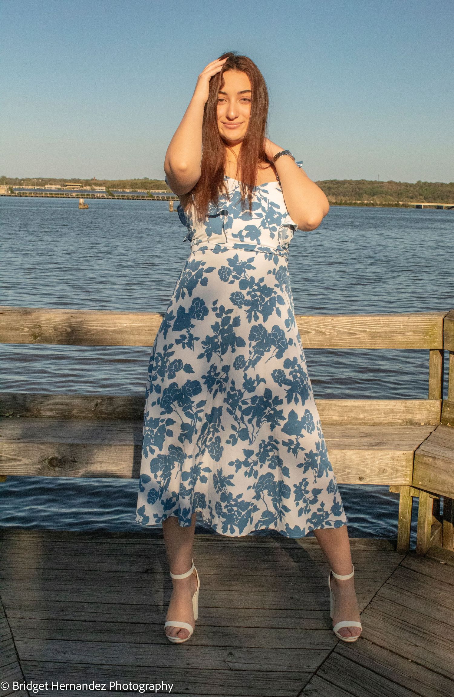 A blue and white floral dress twirls on a wooden deck overlooking a serene waterfront.
