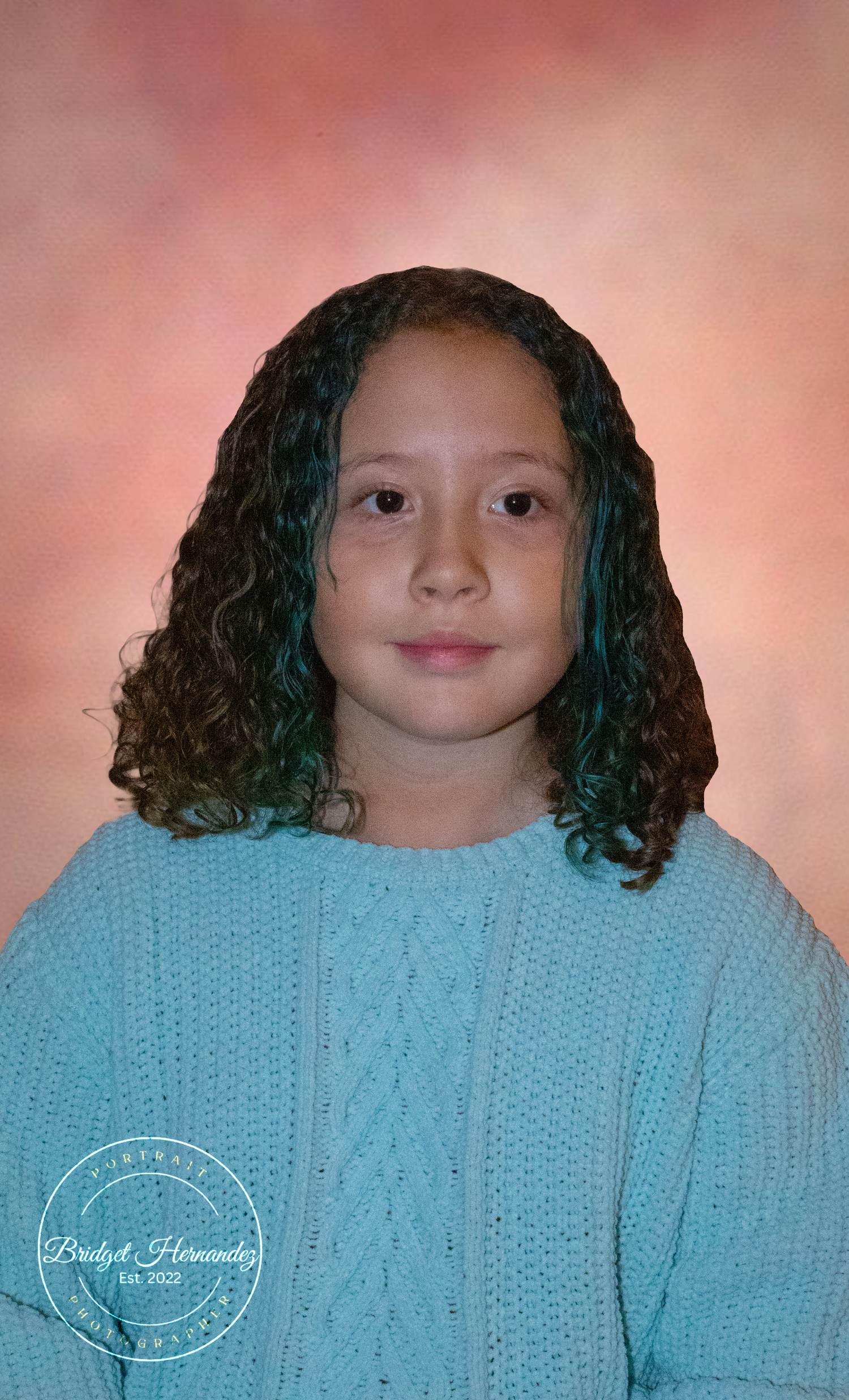 Portrait photo against pink backdrop showing a young child wearing a blue cable knit sweater with curly hair.