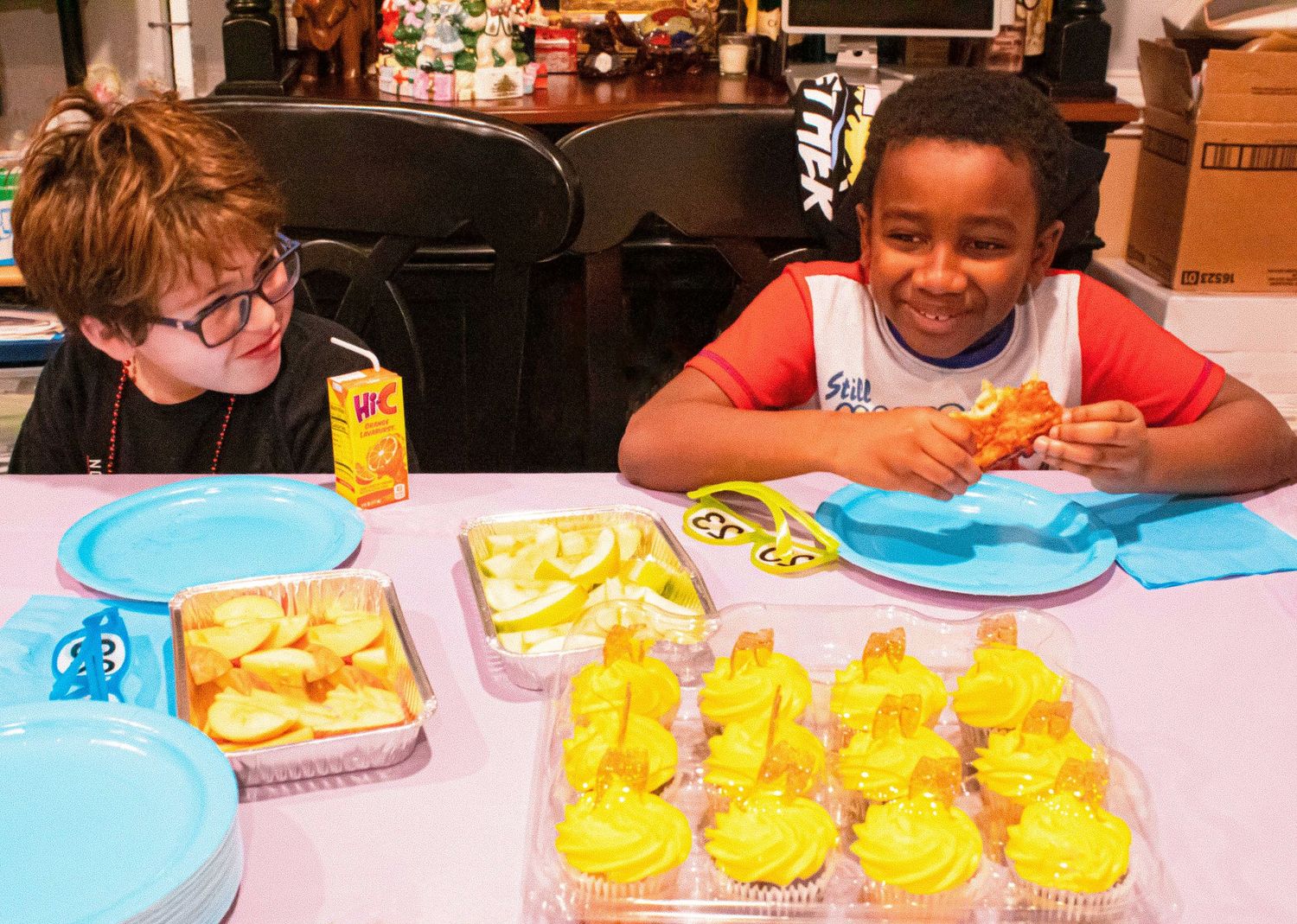 Two children smiling and enjoying snacks at a party table with yellow cupcakes.