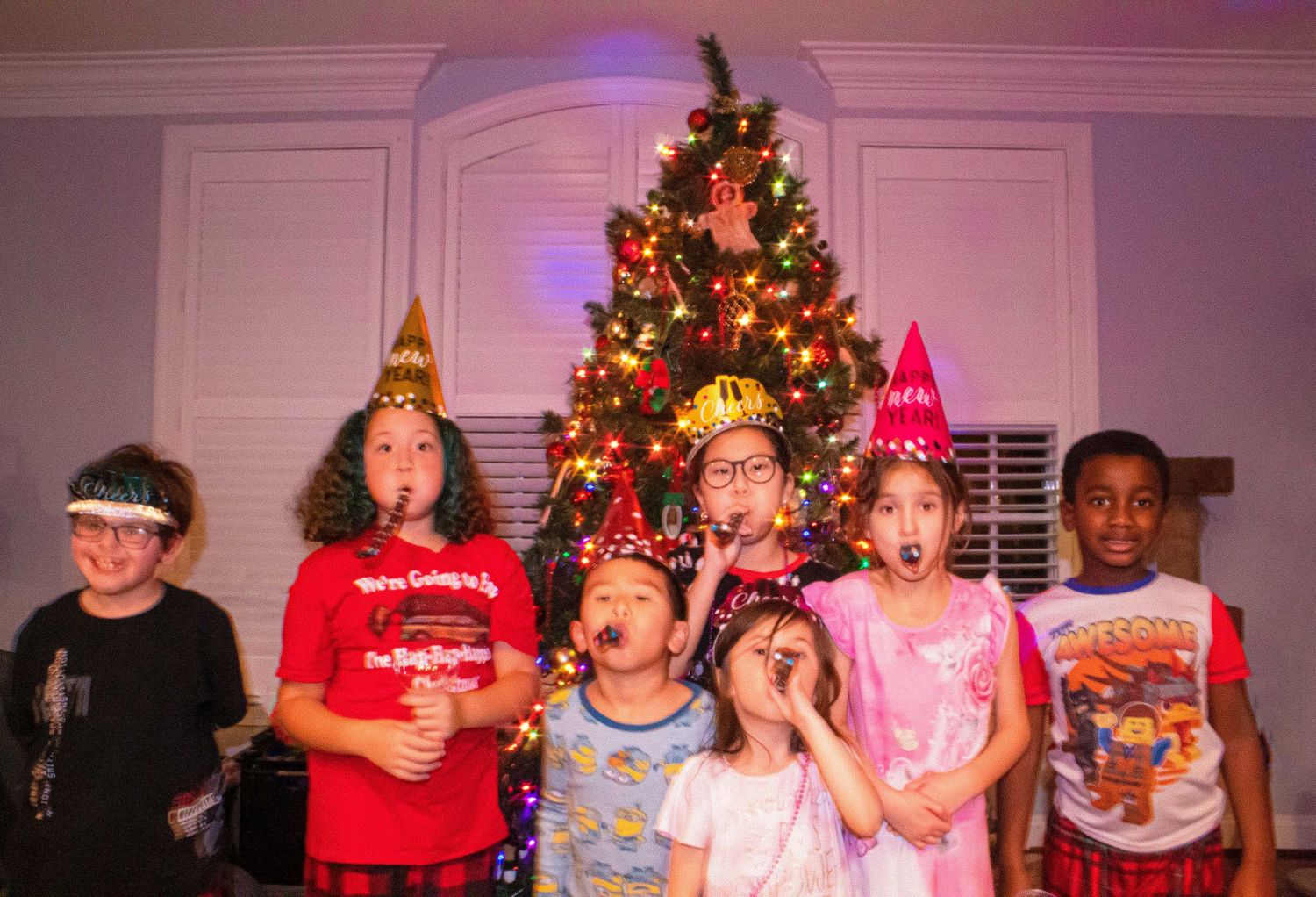 Kids lined up in front of a Christmas tree wearing party hats and pajamas.