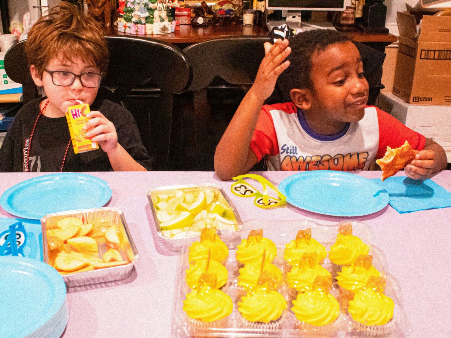 Two children enjoying drinks and snacks at a party with yellow cupcakes on the table.