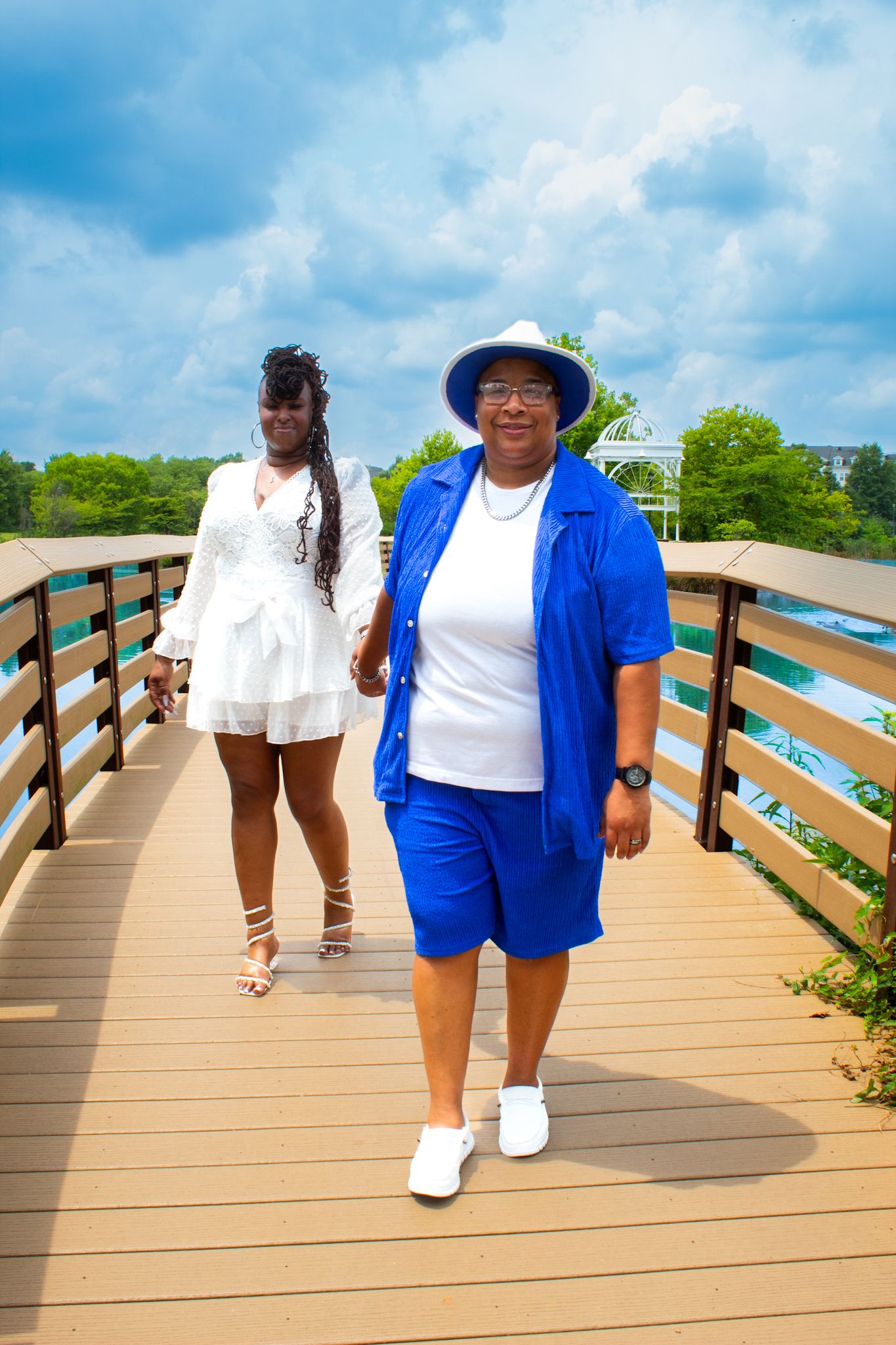 Two people walking on wooden boardwalk wearing blue and white outfits near coastal waters.