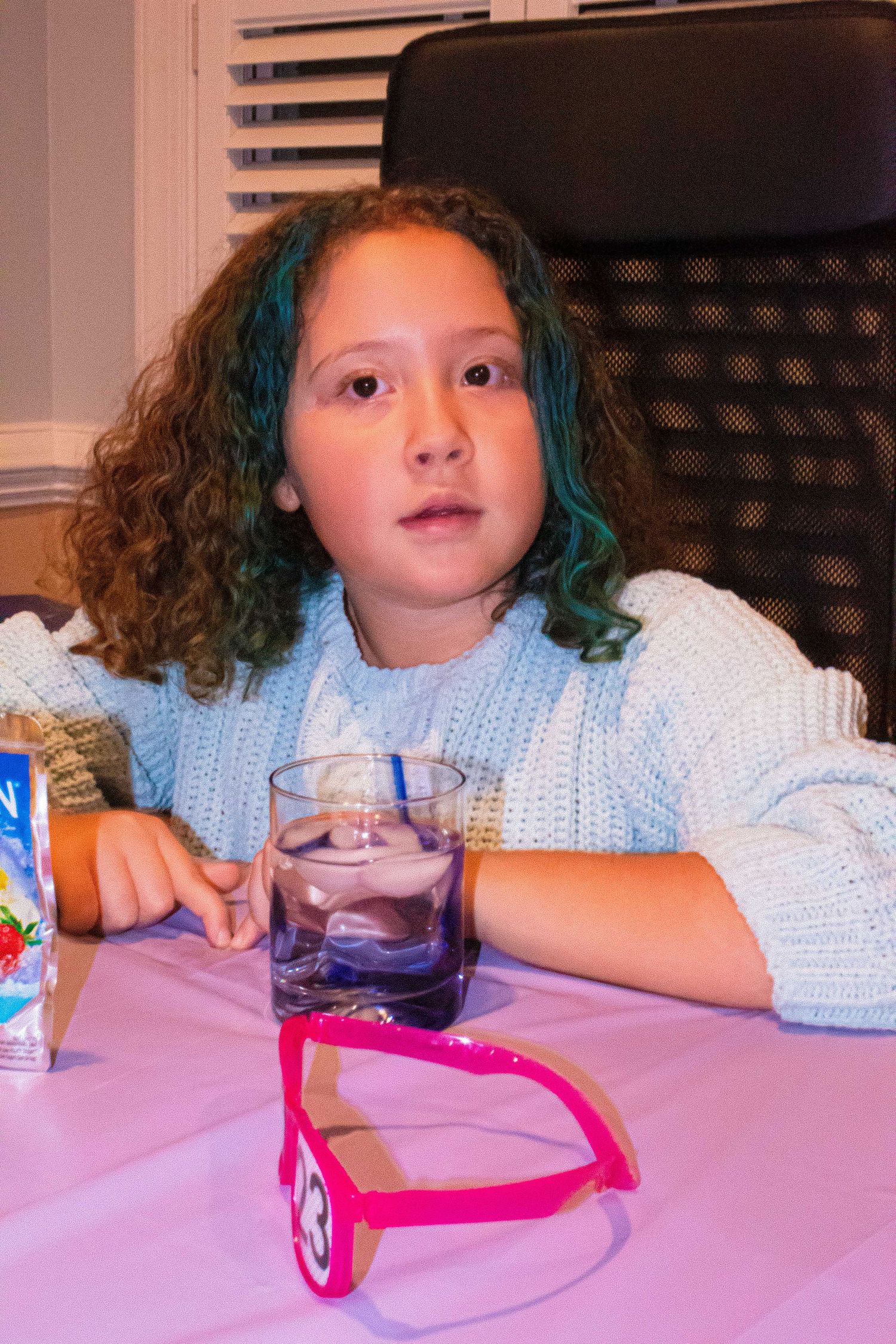 Child in light blue sweater sits at a table with a glass of water and party decorations.