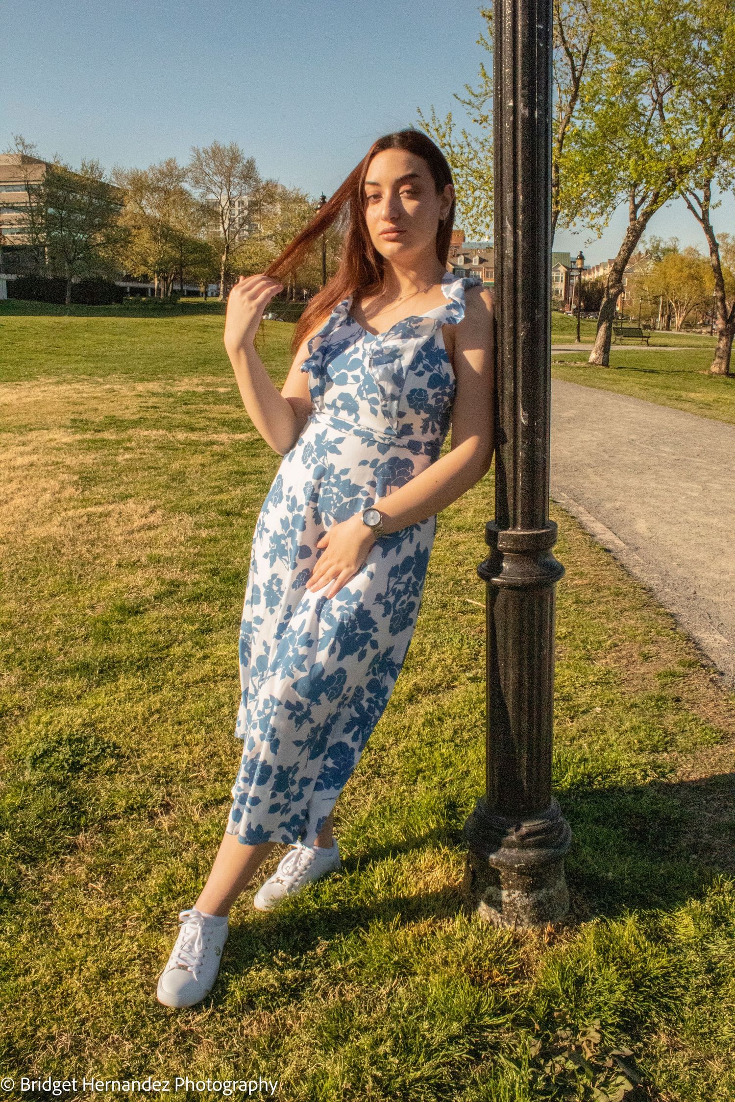 Someone in a blue and white floral dress poses playfully against a lamppost in a park setting on a sunny day.