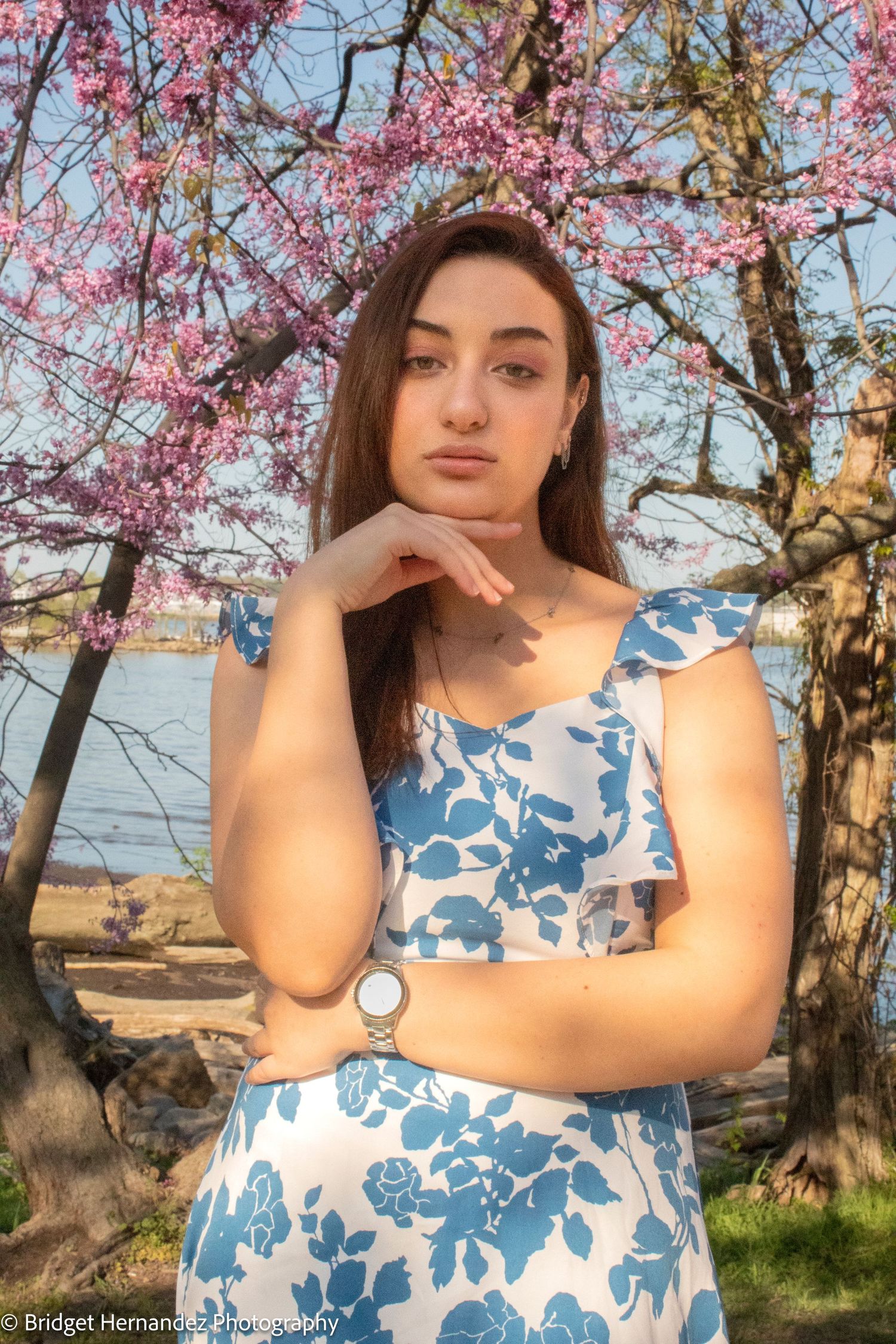 Portrait in floral dress under blooming cherry trees with waterfront view.