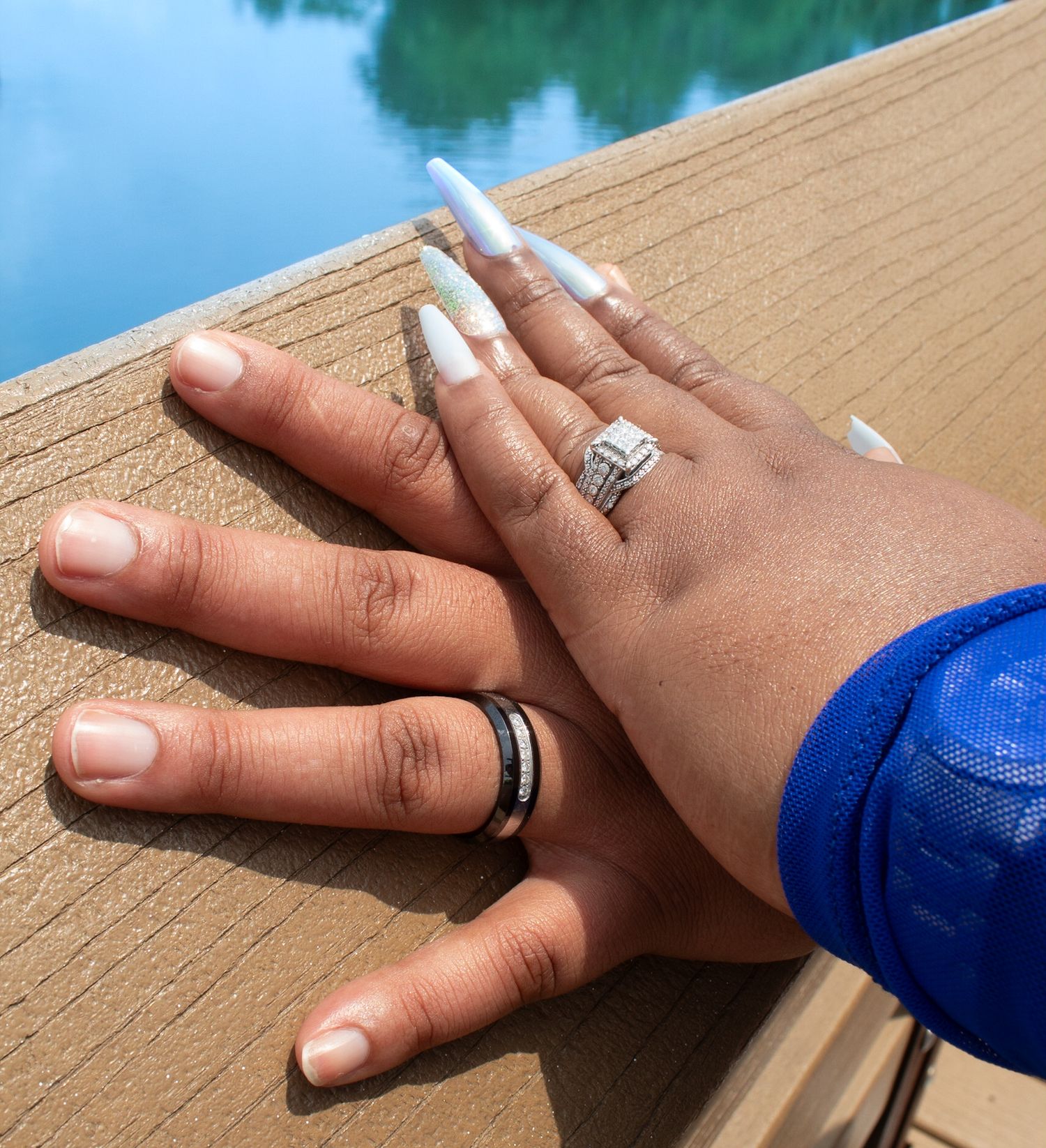 Hands with engagement rings and matching bands displayed on wooden deck railing.