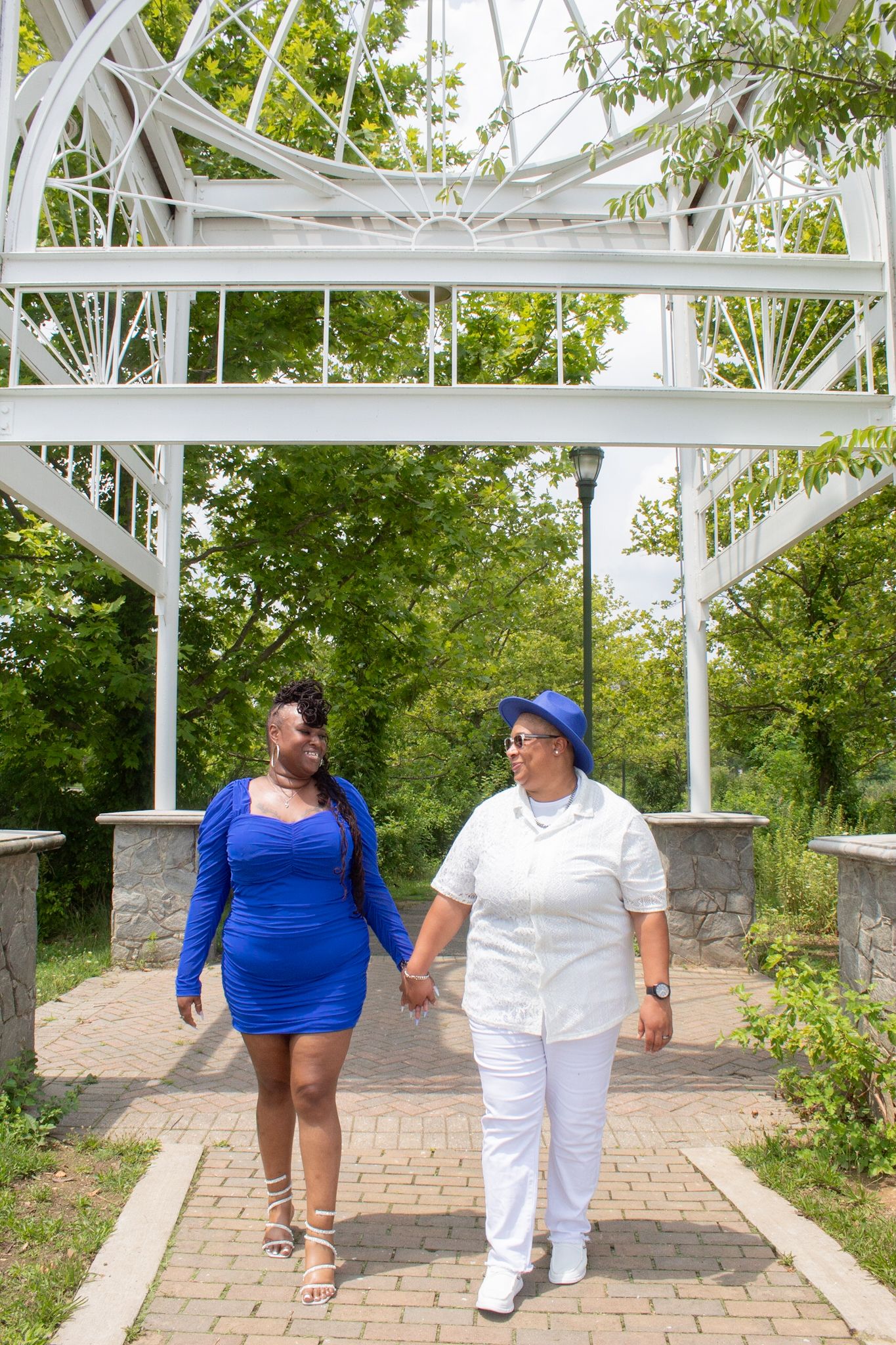 Couple walking together under white garden arbor on brick path surrounded by greenery.
