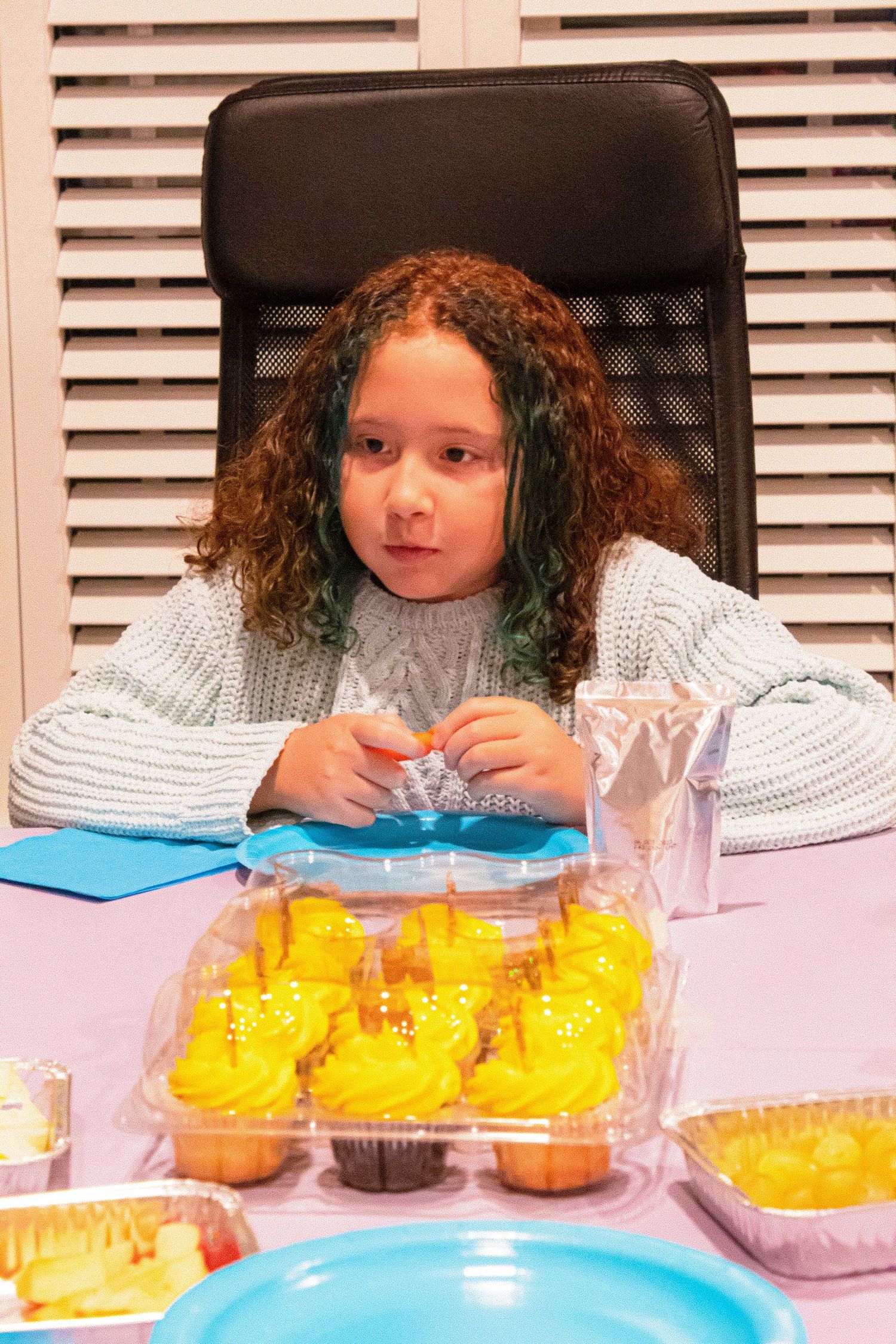 A child in a white sweater sits at a table with yellow cupcakes and blue plates.