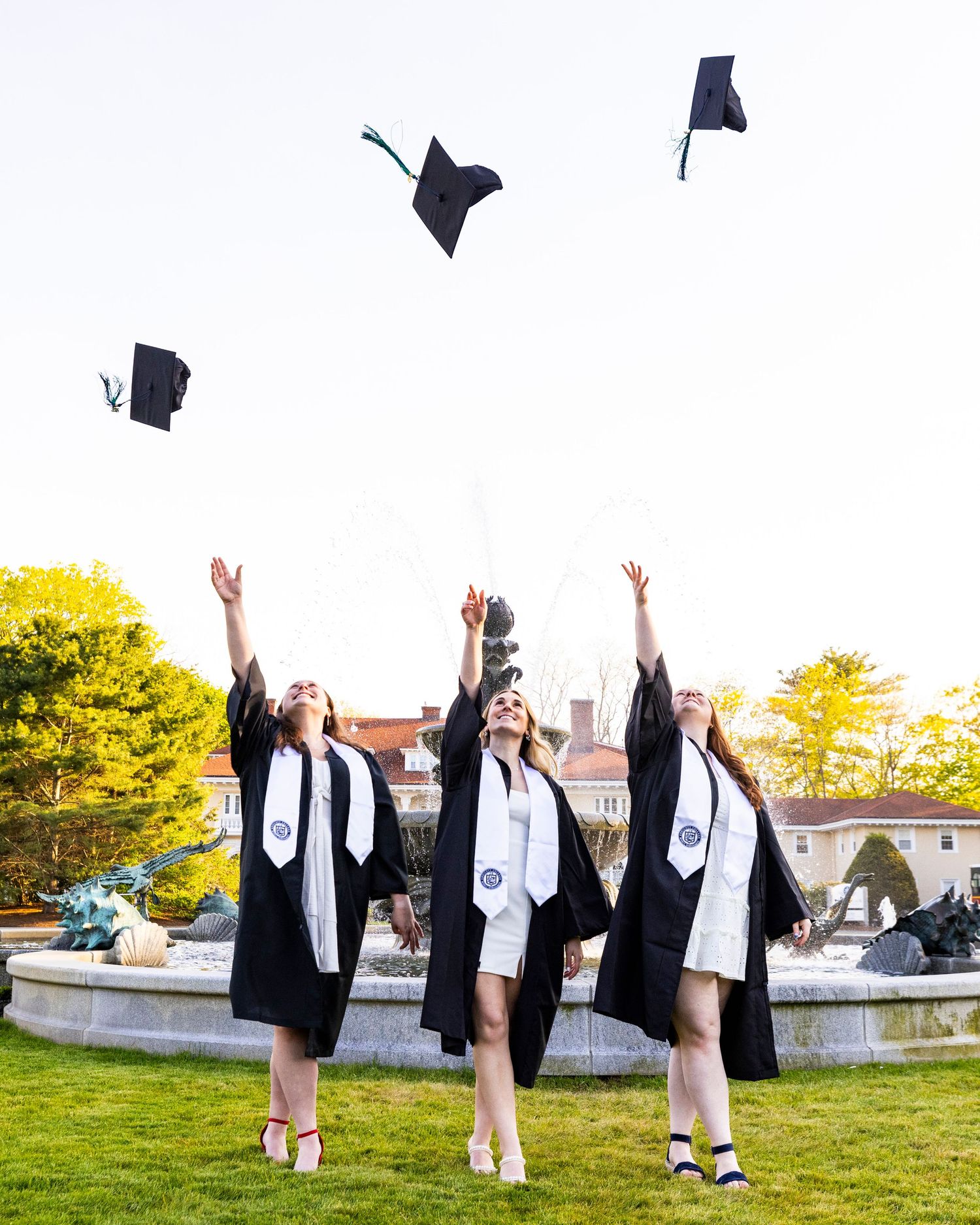 Three female graduates in black robes tossing caps near a fountain during outdoor graduation celebration.